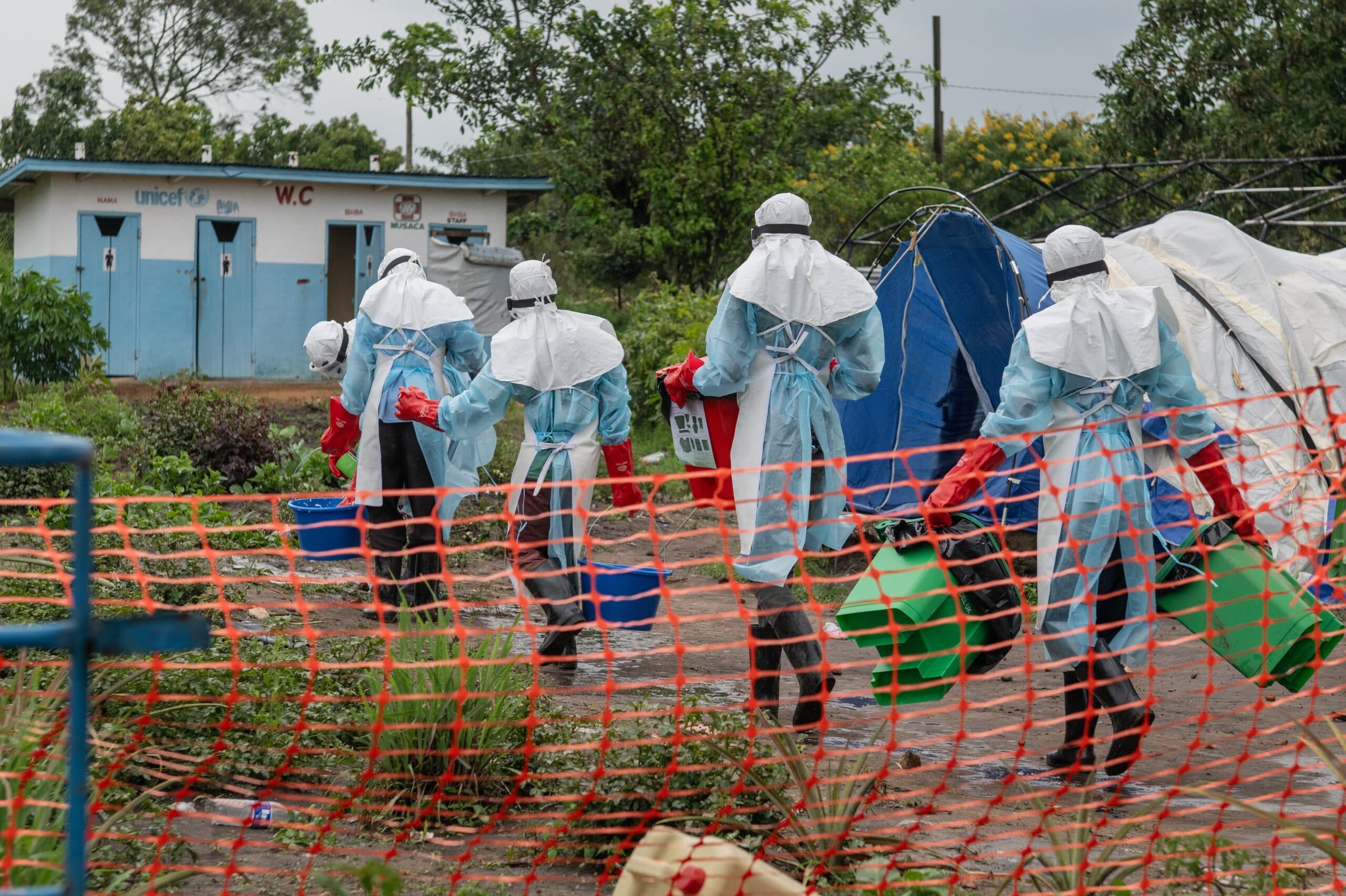 Healthcare workers in protective gear carry supplies at an outdoor facility with tents and orange safety fencing.