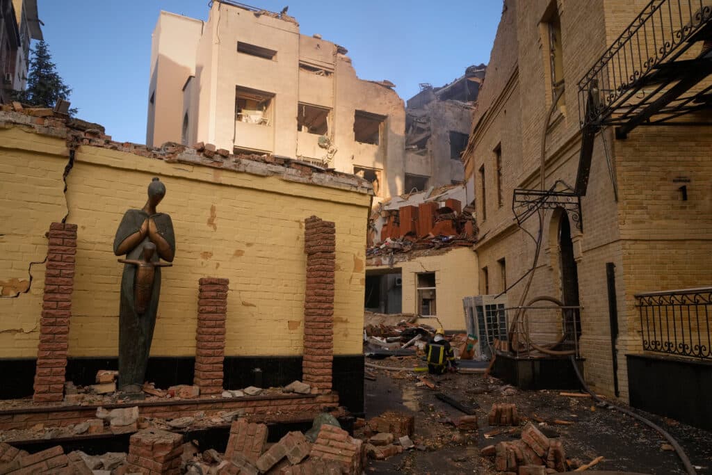 A damaged building surrounded by rubble, with a standing statue and a person in protective gear in the background.