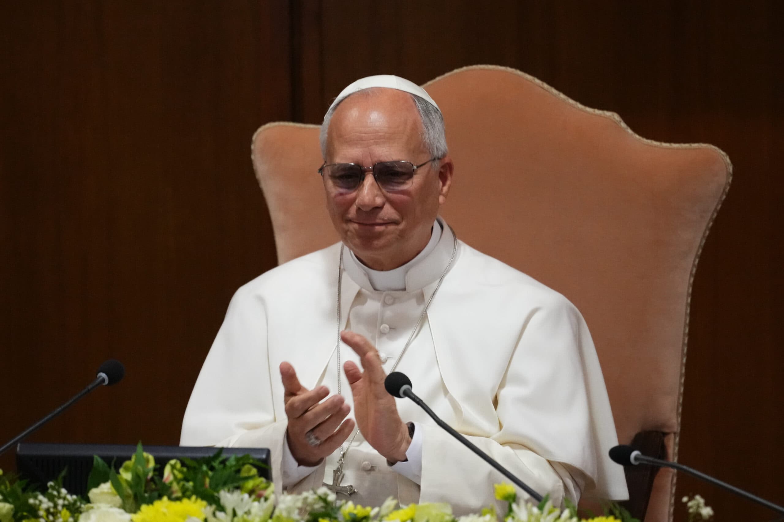 A person in religious attire, wearing a white robe and cap, claps while seated in an ornate chair with flowers in the foreground.
