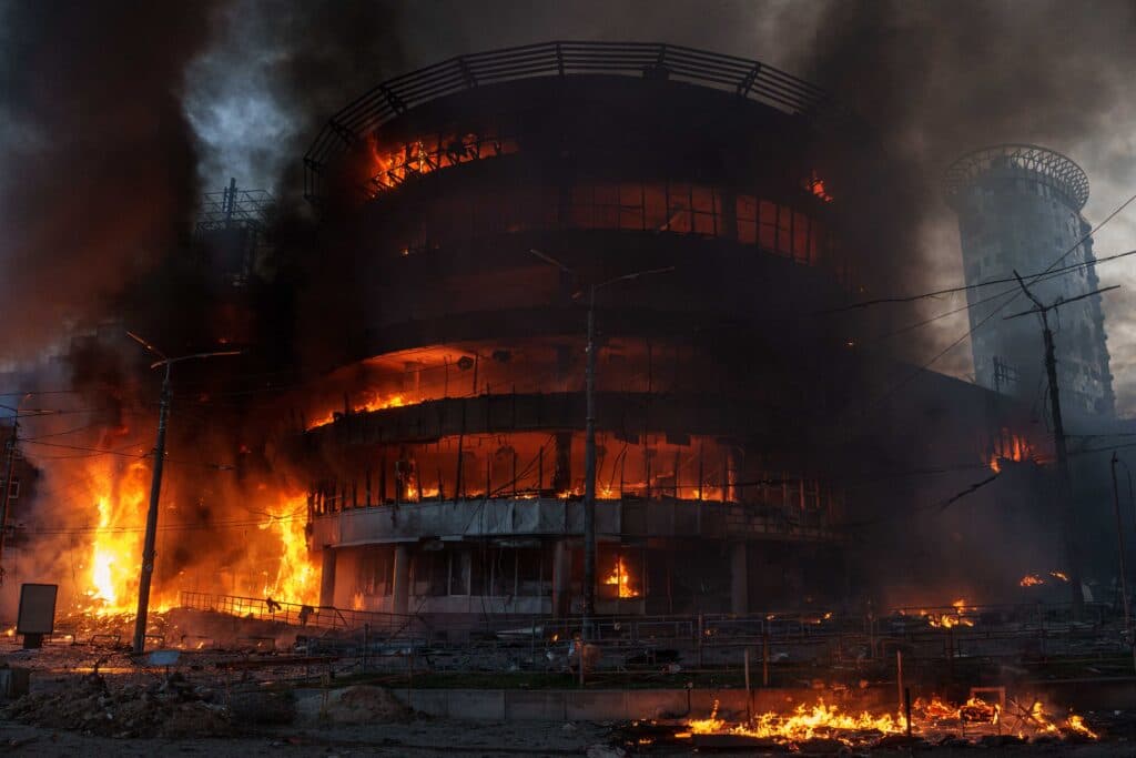 A large building engulfed in flames and smoke against a backdrop of dark skies, surrounded by debris and fire.