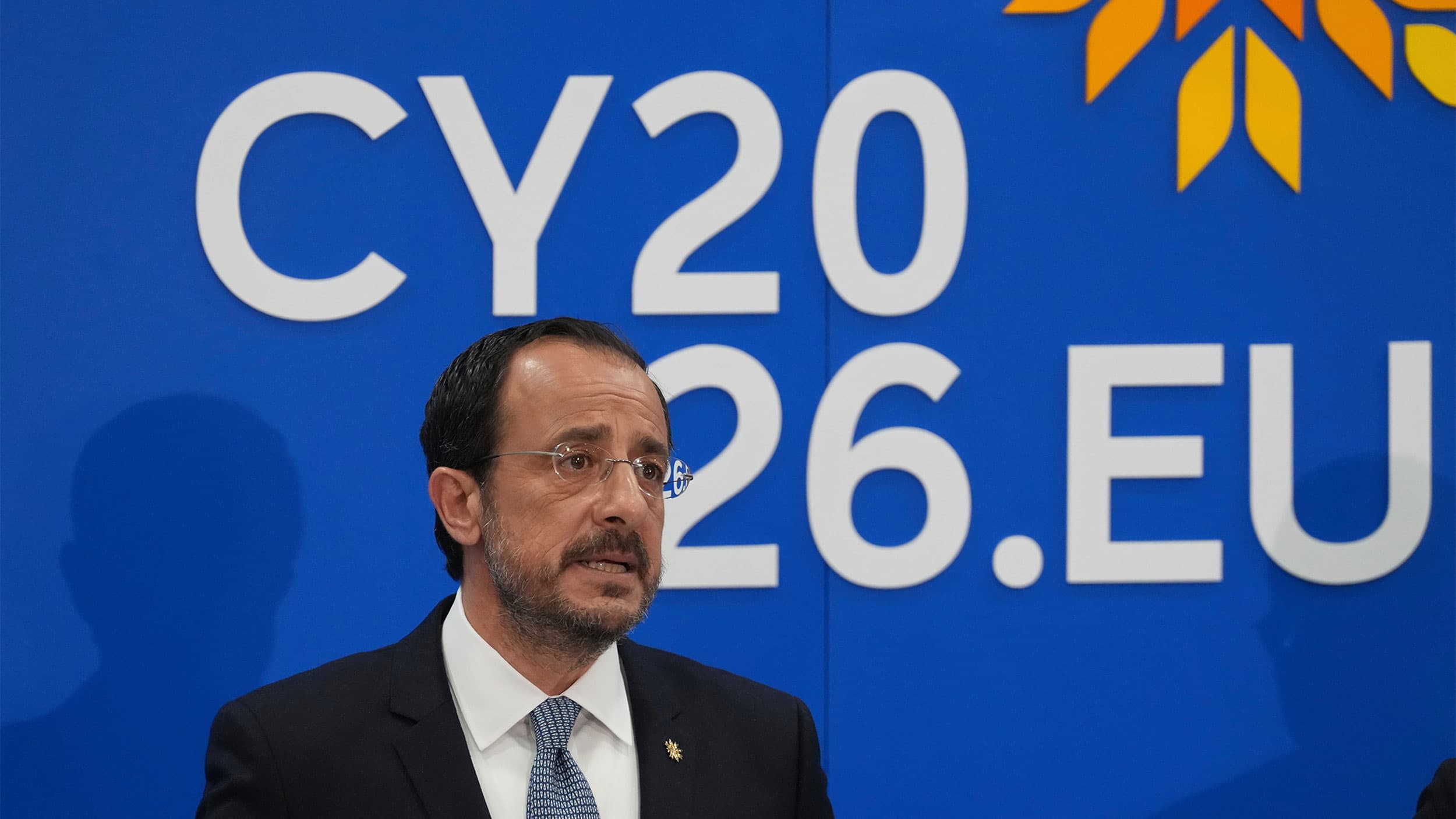 A man in a suit speaks in front of a blue backdrop with "CY2026.EU" text and a yellow emblem.
