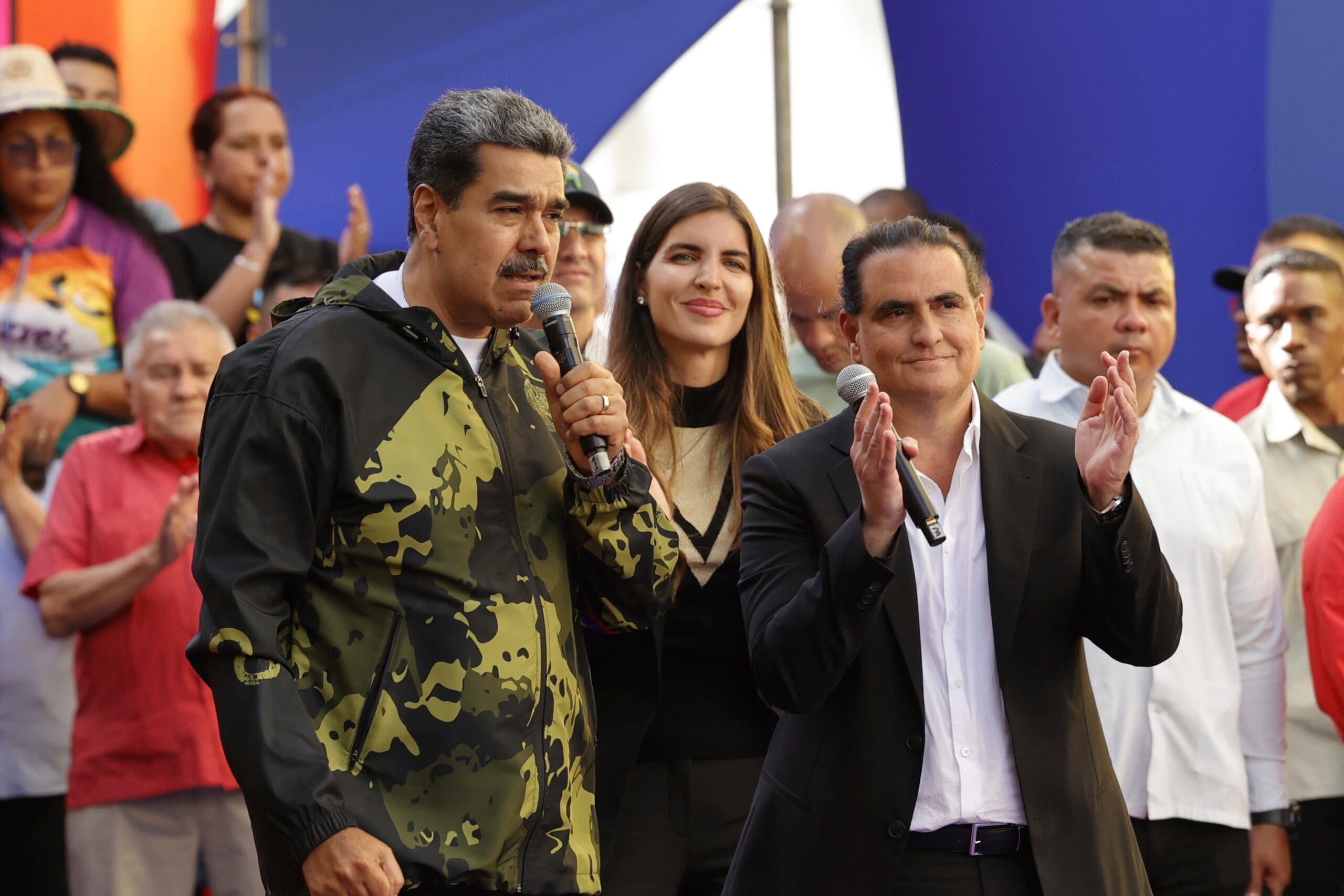 A group of people in formal and casual clothing are gathered, with one man speaking into a microphone and others applauding.