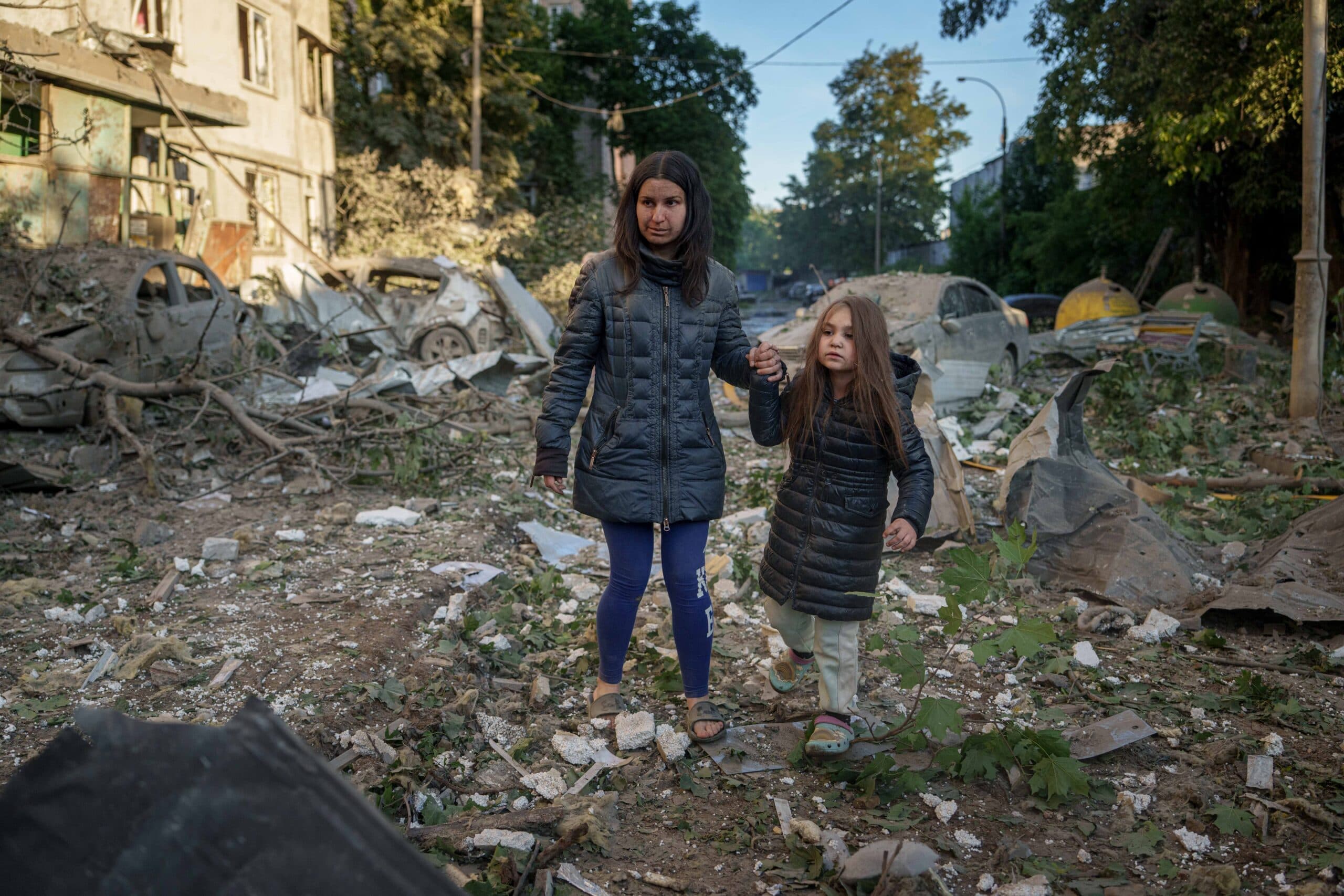 A woman and a child walk hand in hand through a street filled with debris and damaged vehicles.