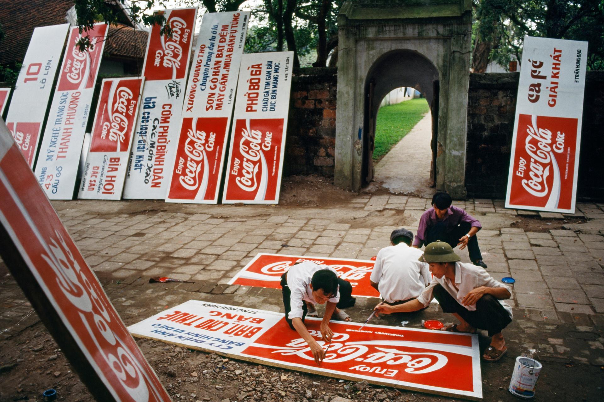 A group of men painting Coca-Cola logos on a dozen signboards. In the 1990s Vietnam revised its constitution to allow the practice of “private capitalism.
