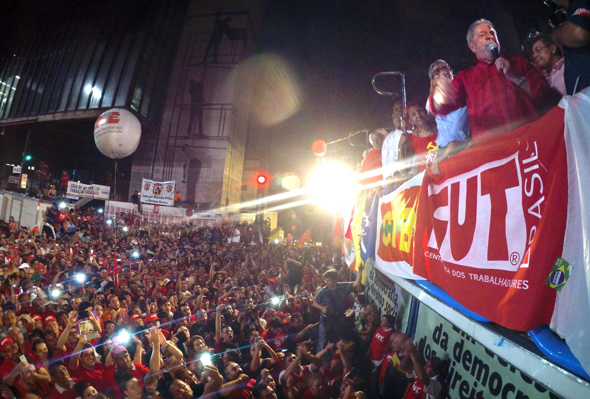 Lula on the side, on raised platform speaking in microphone with crowd below