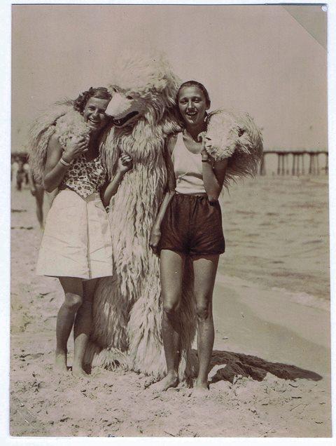 The bear has his arms around two women at the beach