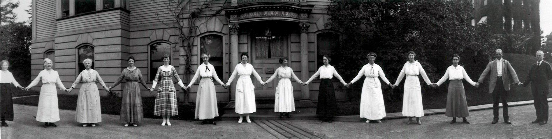 A dozen women hold hands in front of a building on the Harvard campus in 1918. They are all women 