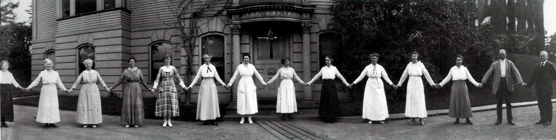 A dozen women hold hands in front of a building on the Harvard campus in 1918. They are all women