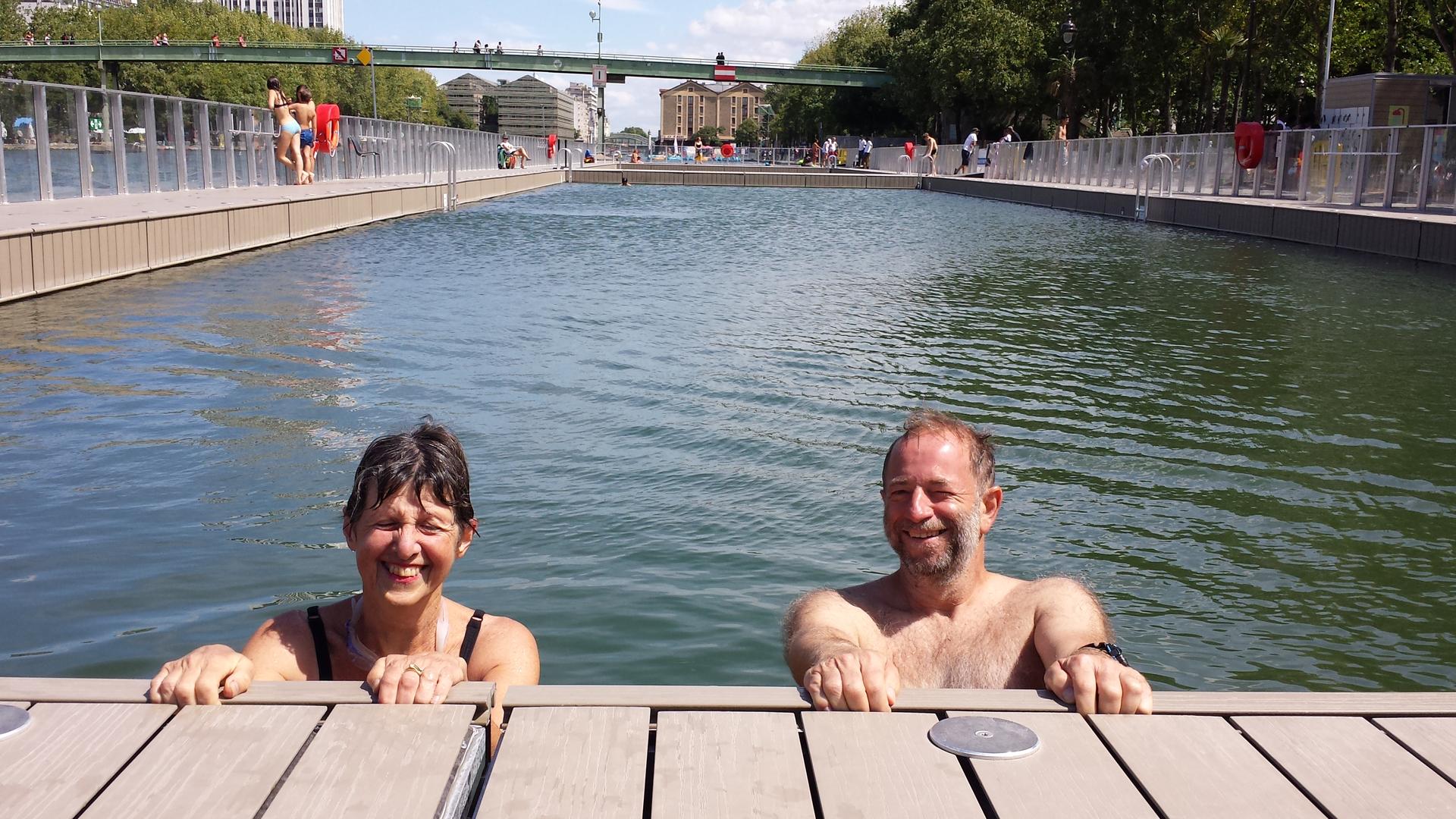 A man and woman beam up at the camera as they hold onto the edge of the pool.