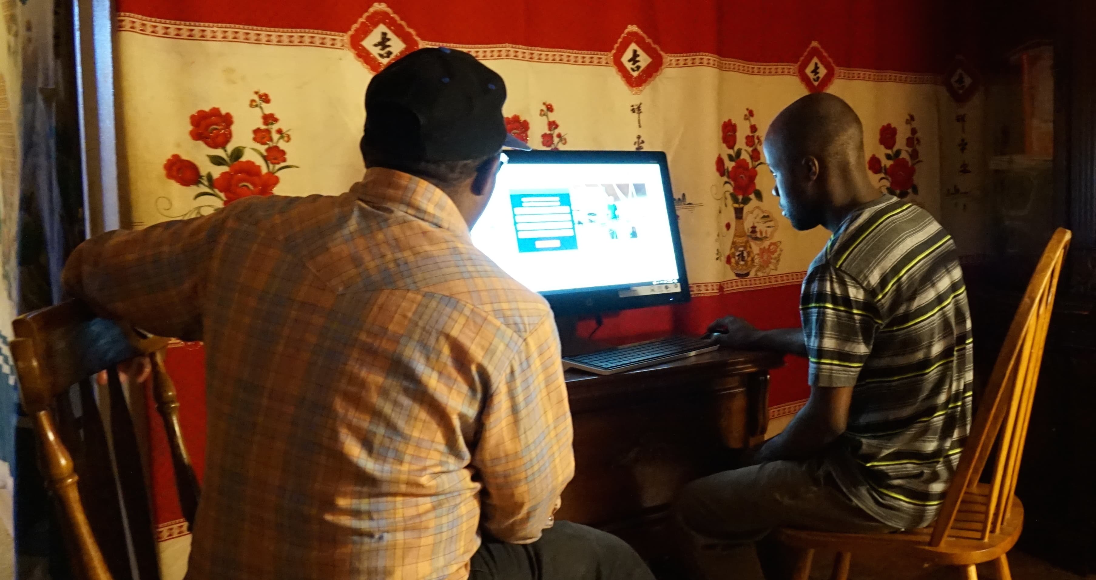 Man sits at computer facing screen with another man sitting next to him looking on