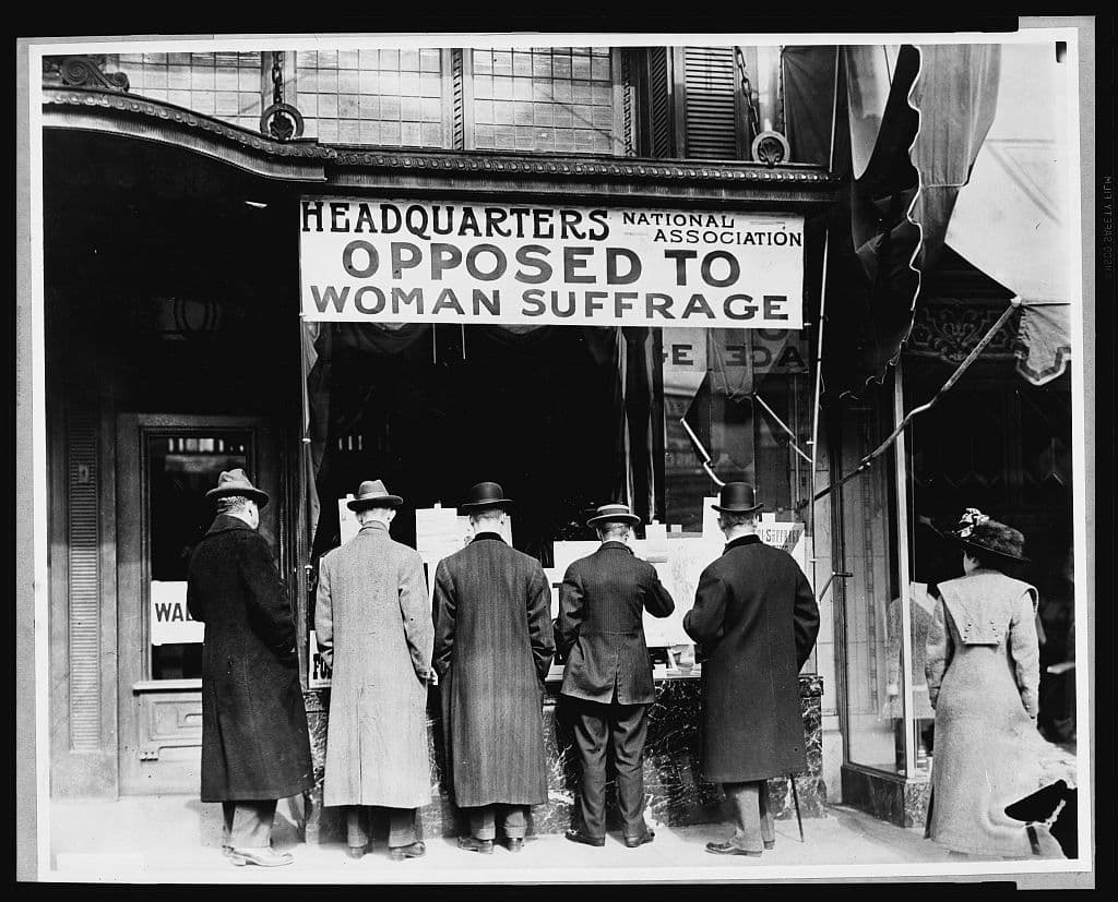 Photograph shows men looking at material posted in the window of the National Anti-Suffrage Association headquarters; sign in window reads