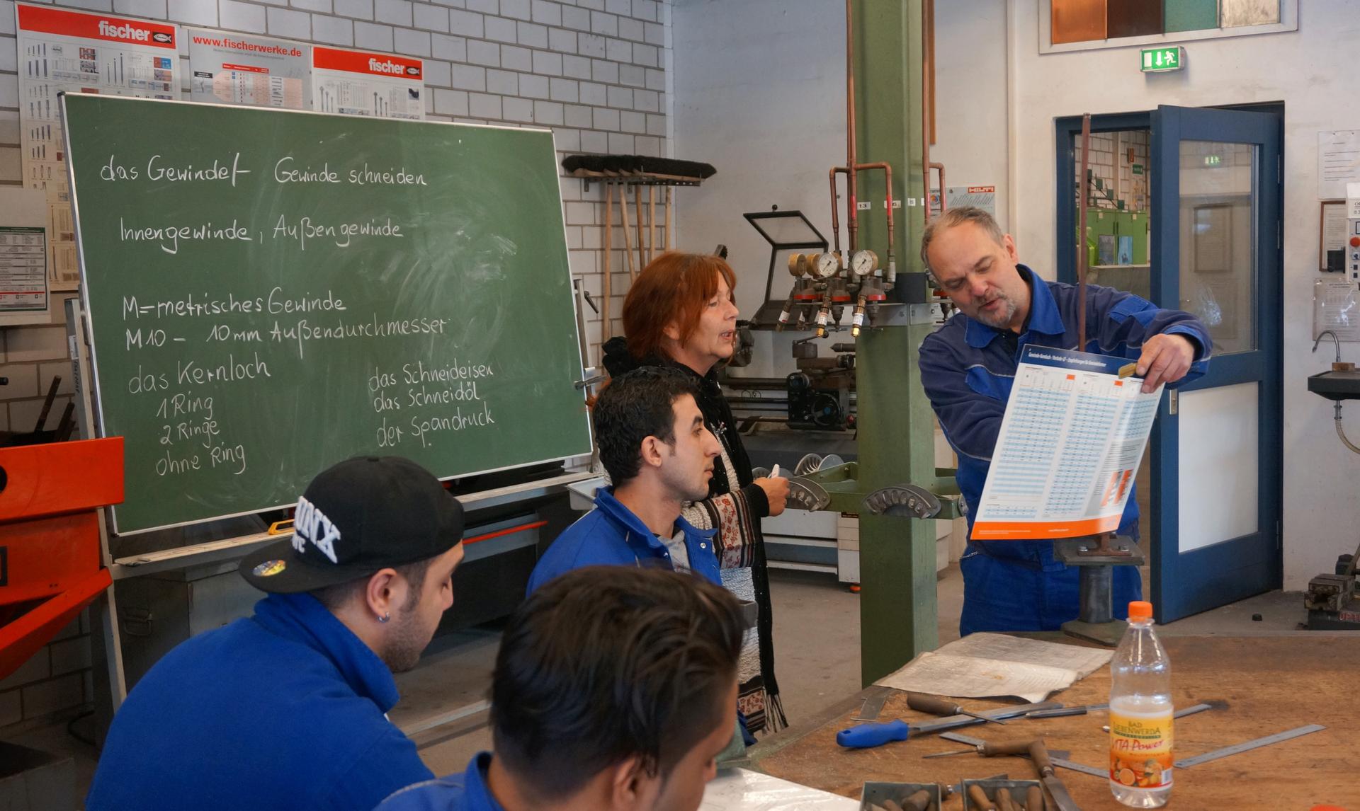 Instructor Gerald Beyer discusses the day's assignment with the class German instructor. As Bayer gives the day's lesson in metalwork, relevant vocabulary is written on the chalkboard. It's a crucial step in getting the refugee participants' language up t