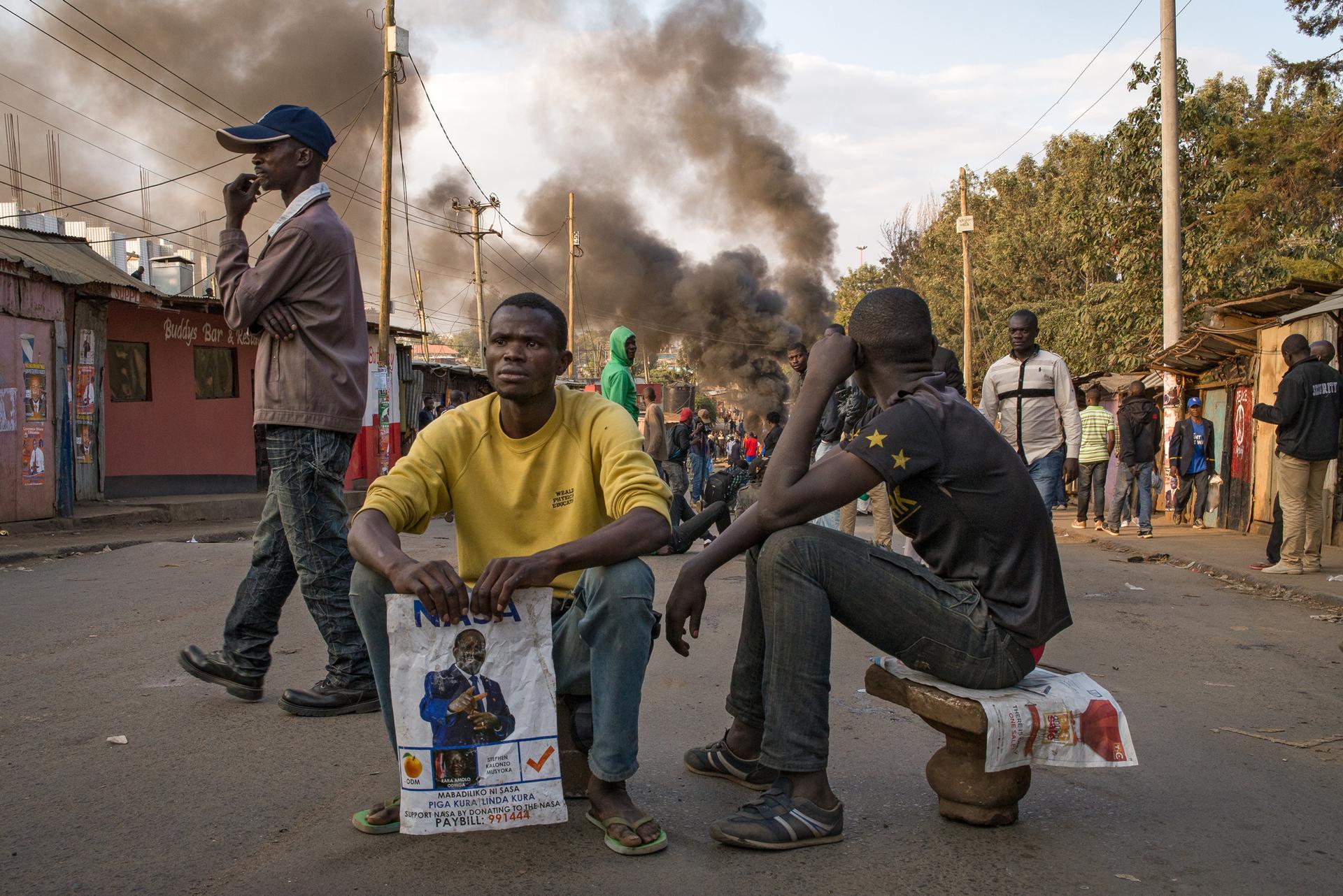 Men watch demonstrators light tires on fire in Nairobi’s informal settlement of Kaibera. The demonstration quickly turned violent as demonstrators armed themselves with stones and clubs bearing nails Wednesday evening.