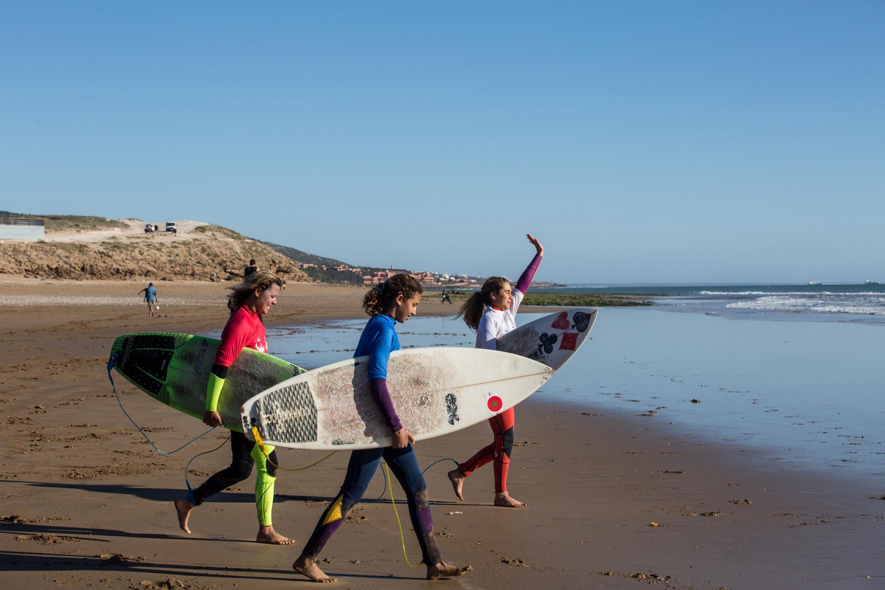 Ghita Guemmi (right), and Fatima Berrada (left), Lilias Tebbaï (center) approach the water with their surfboards.