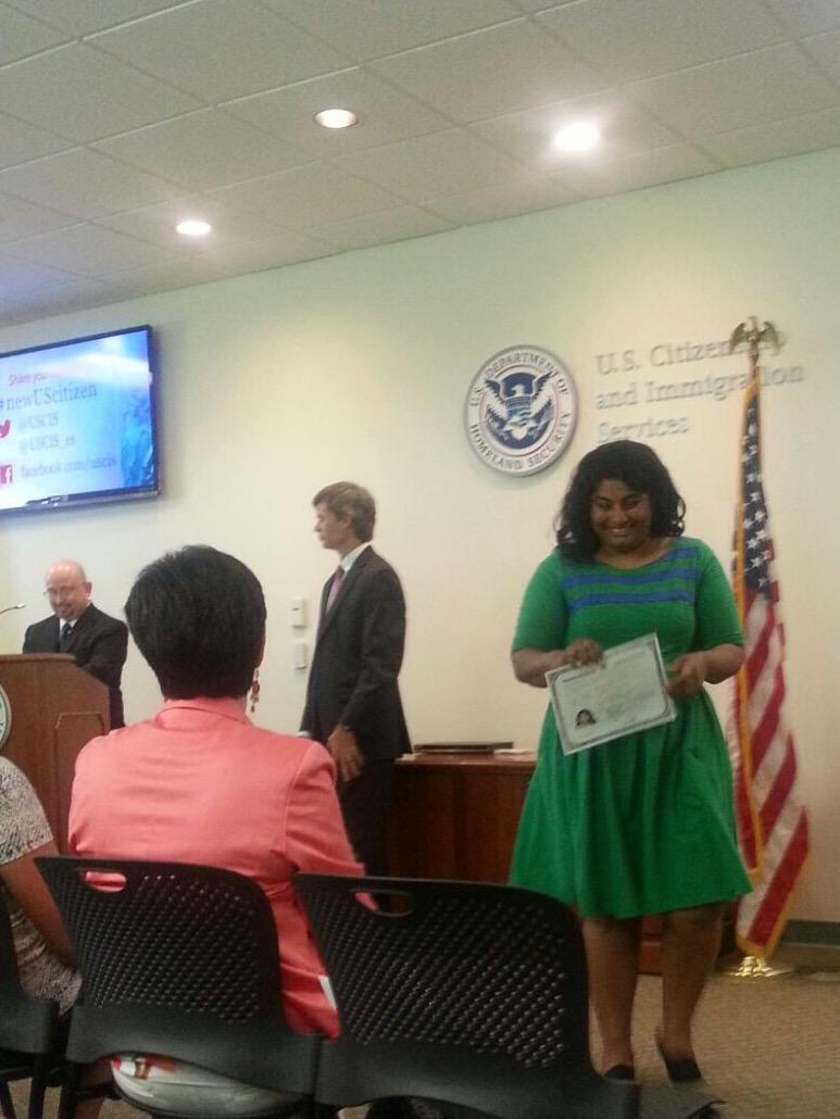 Blurry image of Koshy in front of USCIS sign and seal, with her citizenship certificate