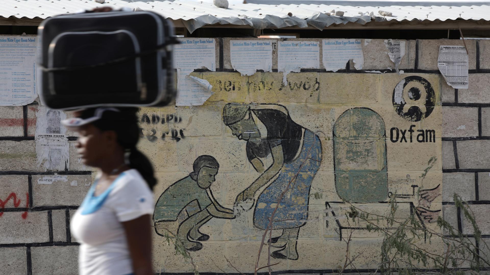 A woman walks carrying a suitcase on her head next to an Oxfam sign in Corail, a camp for displaced people of the 2010 earthquake, on the outskirts of Port-au-Prince, Haiti, Feb. 13, 2018.