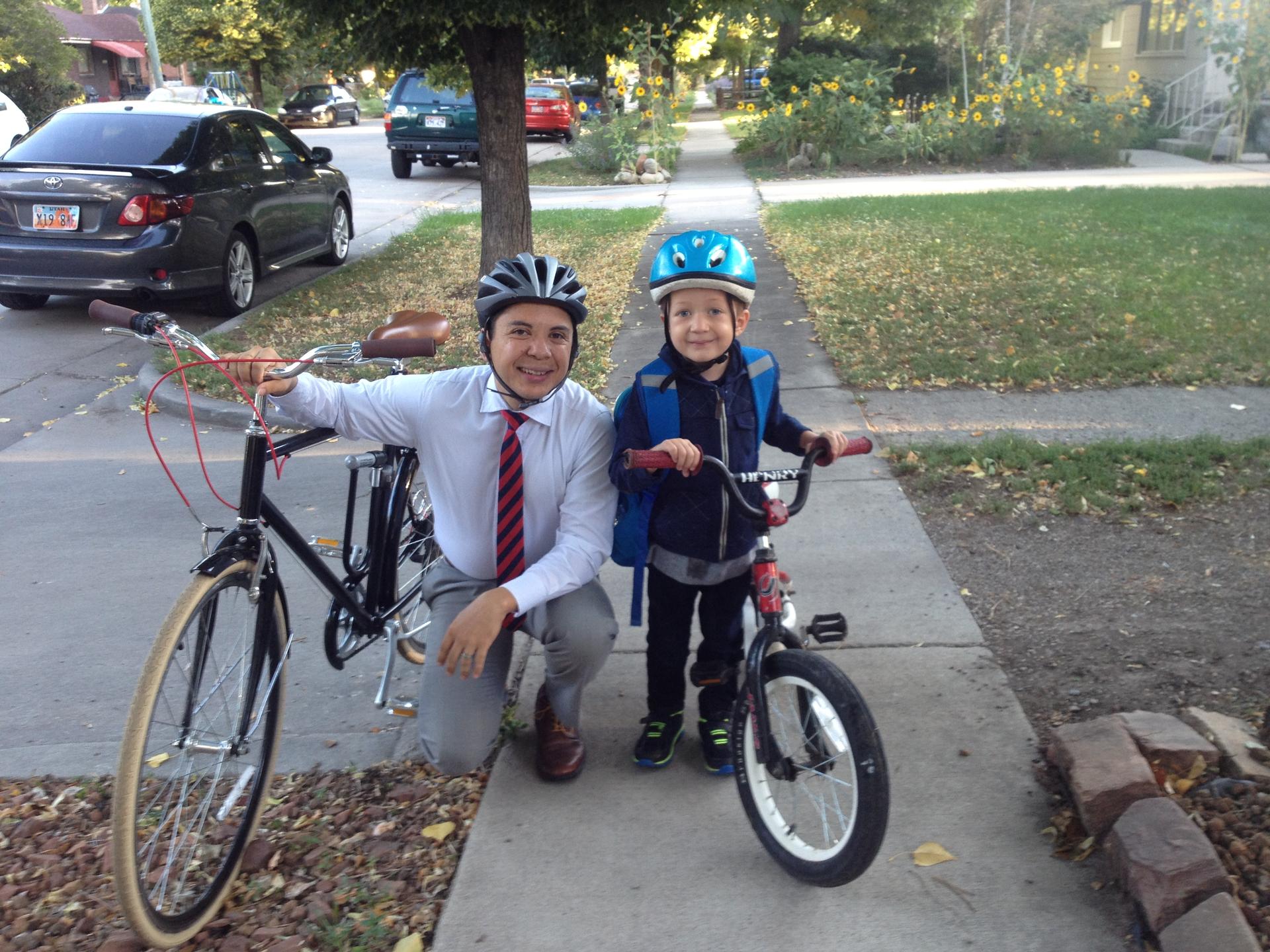 Man wearing helmet and tie kneels on sidewalk next to boy on bike, also wearing helmet