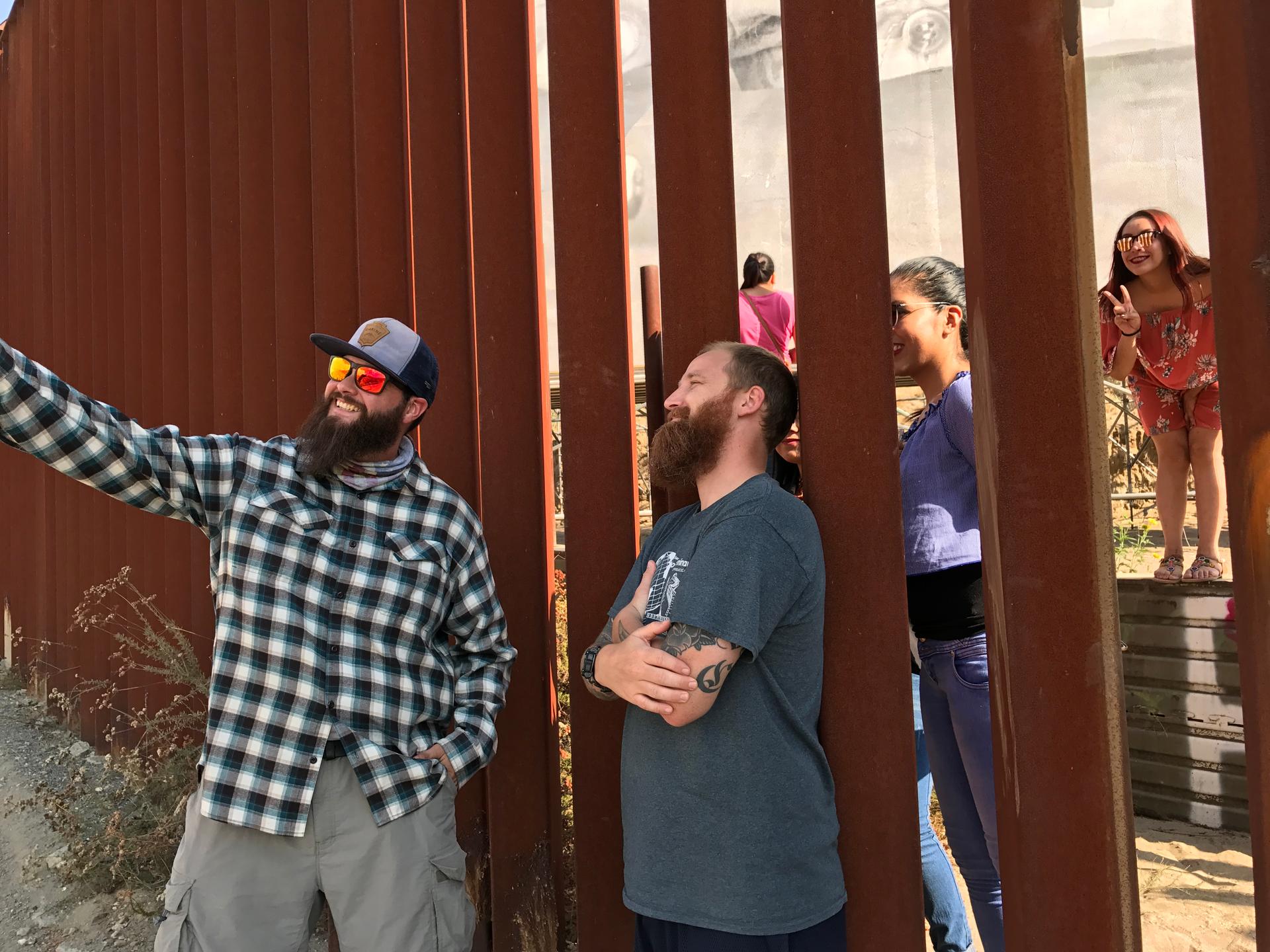 Two smiling men in beards hold up a phone as they stand in front of fence; on other side, you can see women smiling and making the peace sign with their fingers