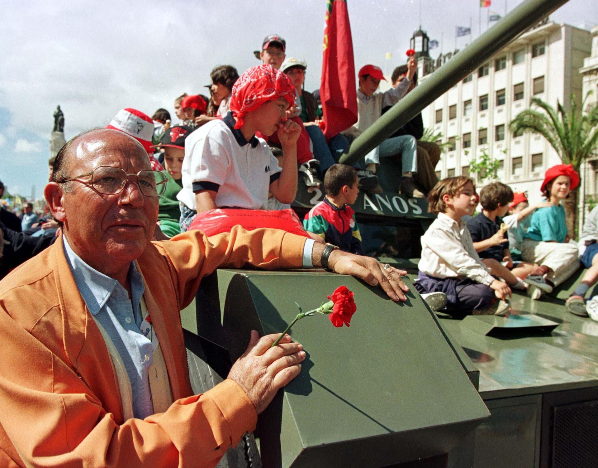 An old Portuguese man holding a carnation flower rides on the side of a carnival armoured car surrounded by children during a parade in Lisbon celebrating the 1974 Carnation Revolution April 25.
