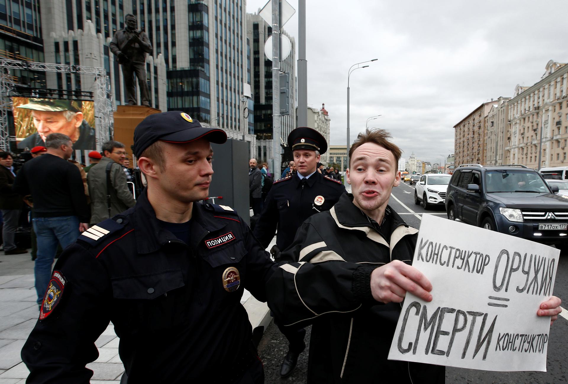 A man is detained by police officers at the unveiling of the Kalashnikov statue on Tuesday. His sign reads: 