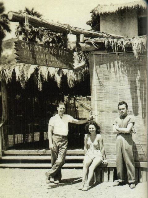 Black and white photo of three people in front of bar entrance, island scene