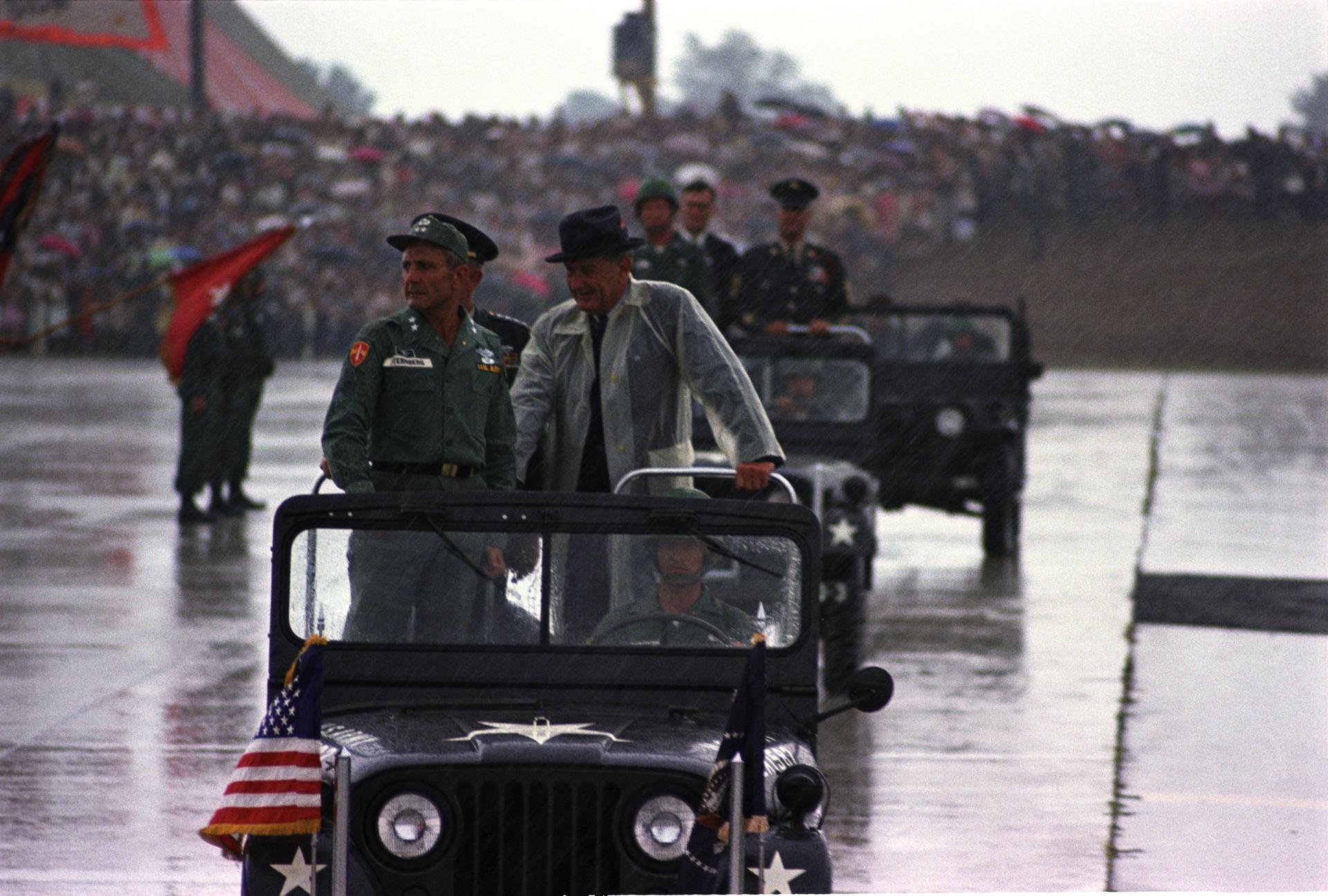 Standing in a jeep during a rainstorm (L-R:) Maj. Gen. Ben Sternberg, Pres. Lyndon B. Johnson 