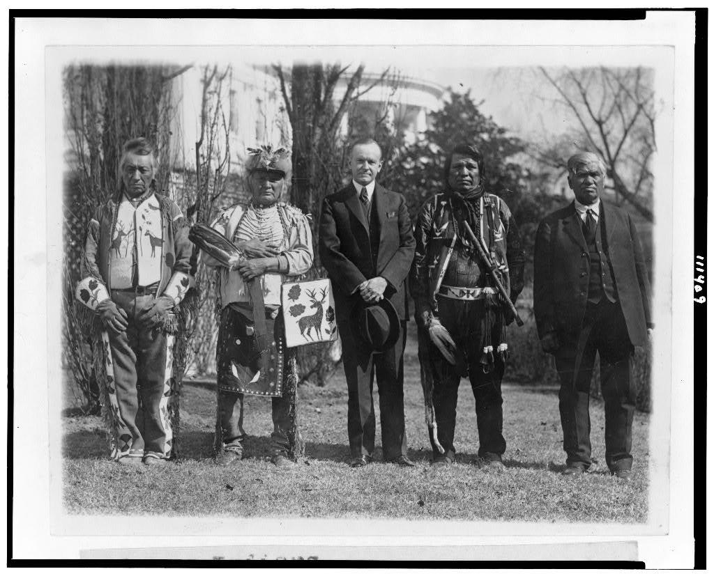 President Calvin Coolidge stands in front of the White House with four Native Americans, one of whom is dressed in suit, tie and vest, and the others wearing traditional dress.