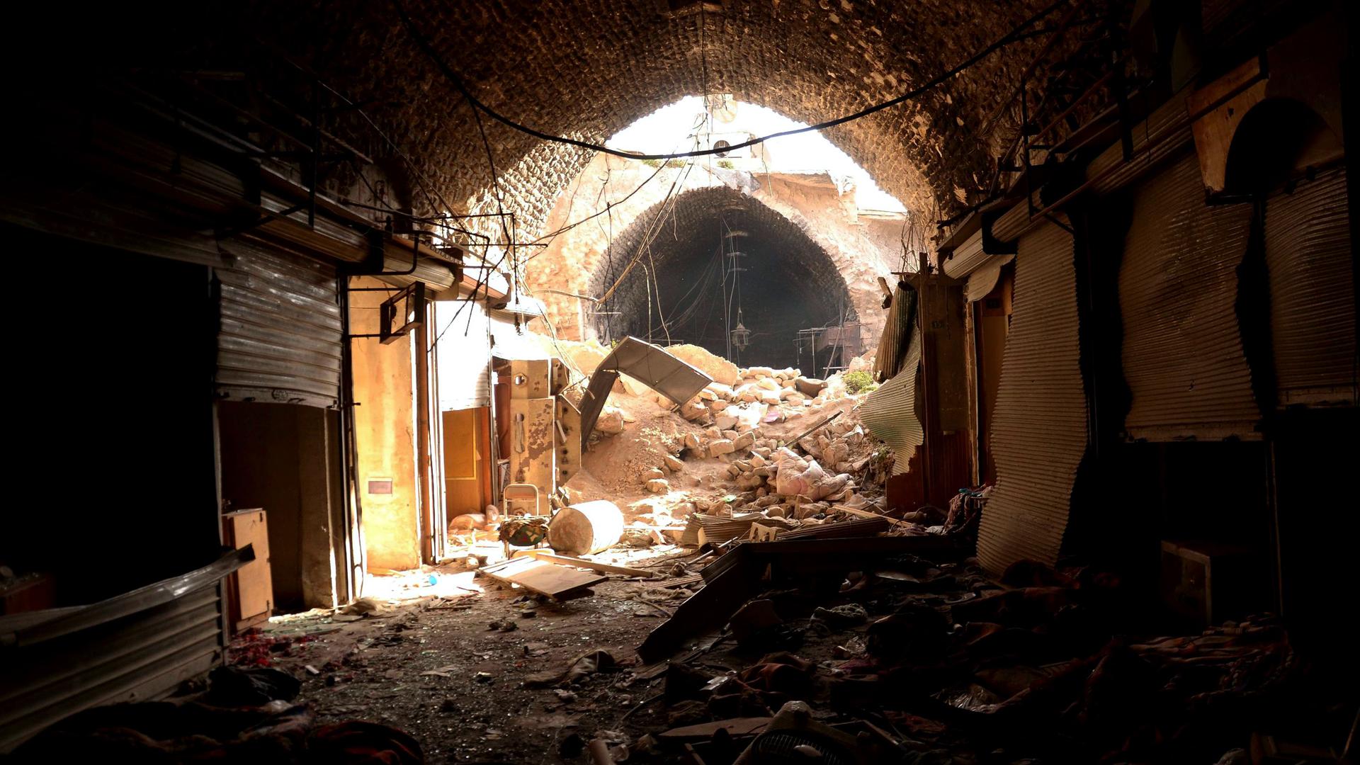 Rubble fills a tunnel that was a souk shop. Some shop locations are visible, with metal doors pulled down. There are no people, no lights.