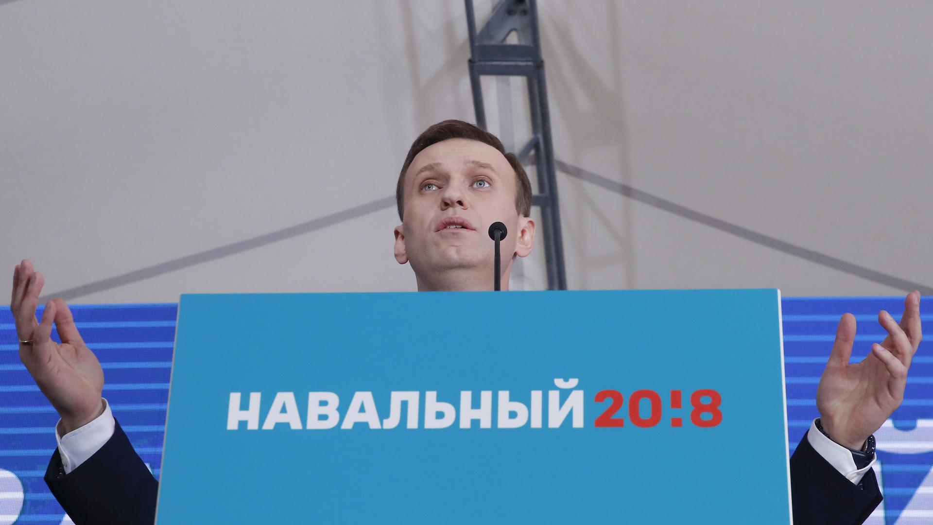 Alexei Navalny speaks behind a podium covered with a blue sign with red lettering in Russian and holds his hands up as he looks out on the crowd.