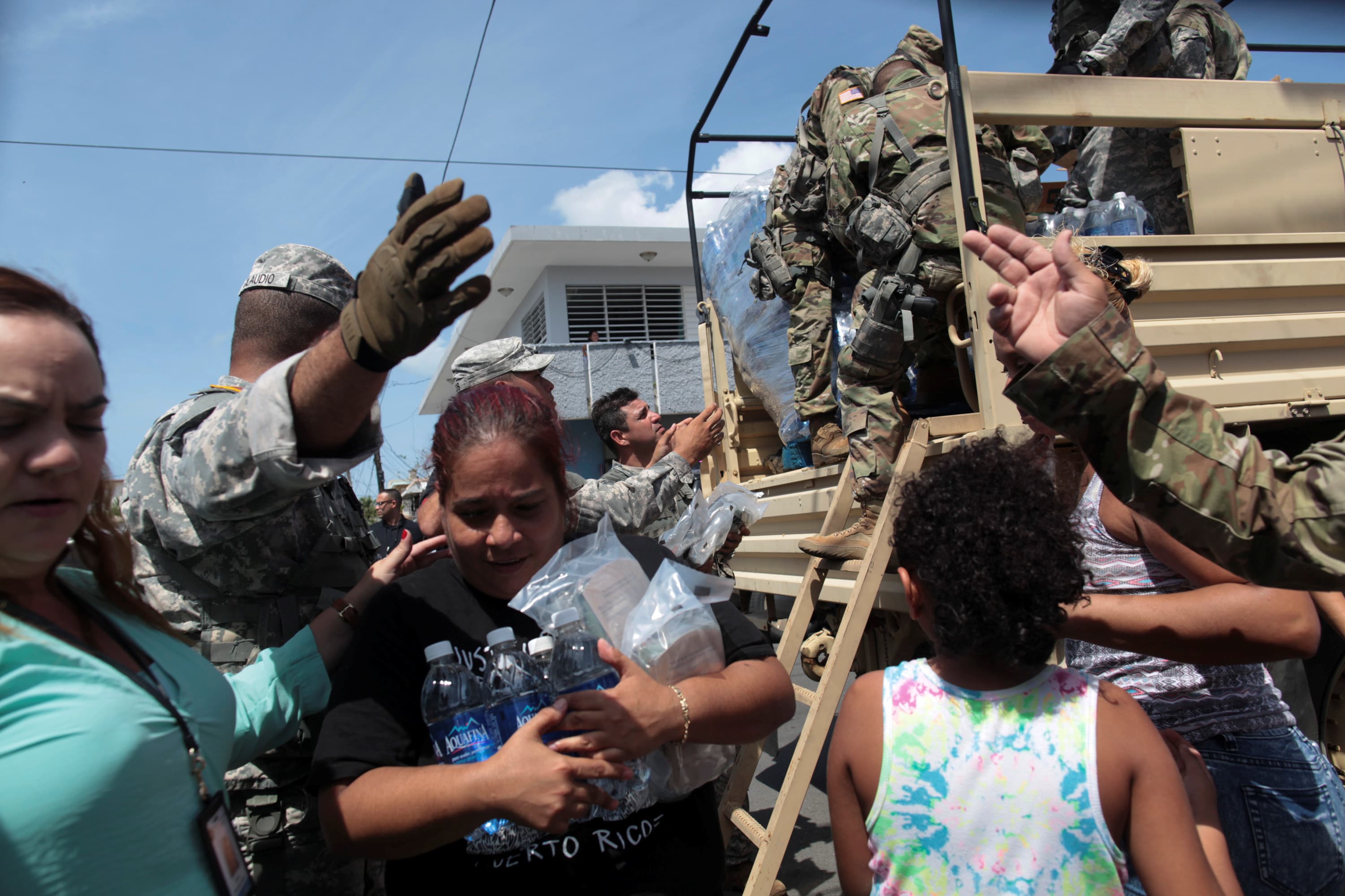 People in uniforms distribute goods off a truck, with woman in the foreground holding water and packages
