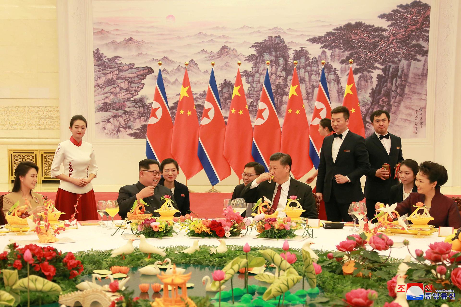 North Korean leader Kim Jong-un and wife Ri Sol Ju, and Chinese President Xi Jinping and wife Peng Liyuan sit at a large round table for dinner and toast each other.
