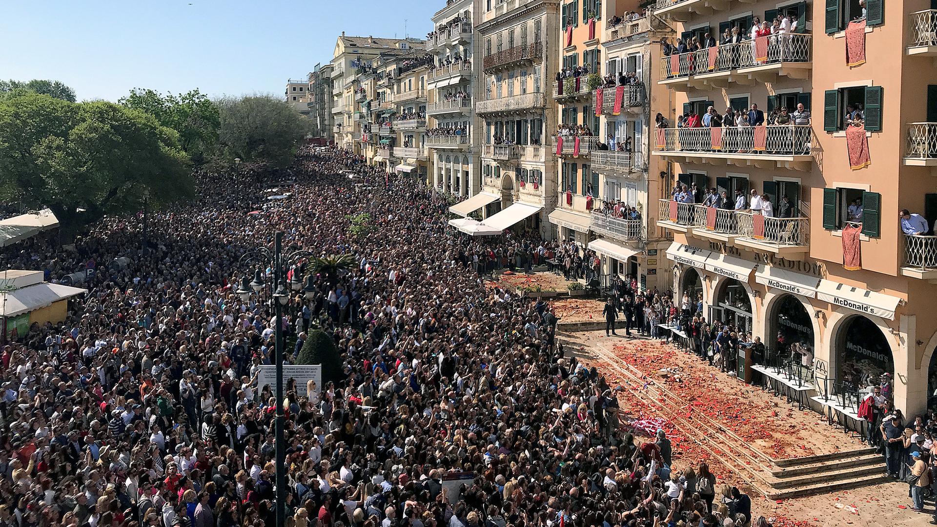 Crowds of people stand in the street while others hang out of windows. Below the windows are the red shards from the clay pots falling on the pavement.