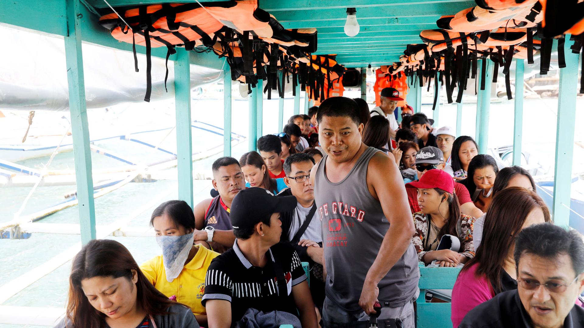 A man walks between seats of a motorboat filled with tourists. Overhead are bright orange life vests.