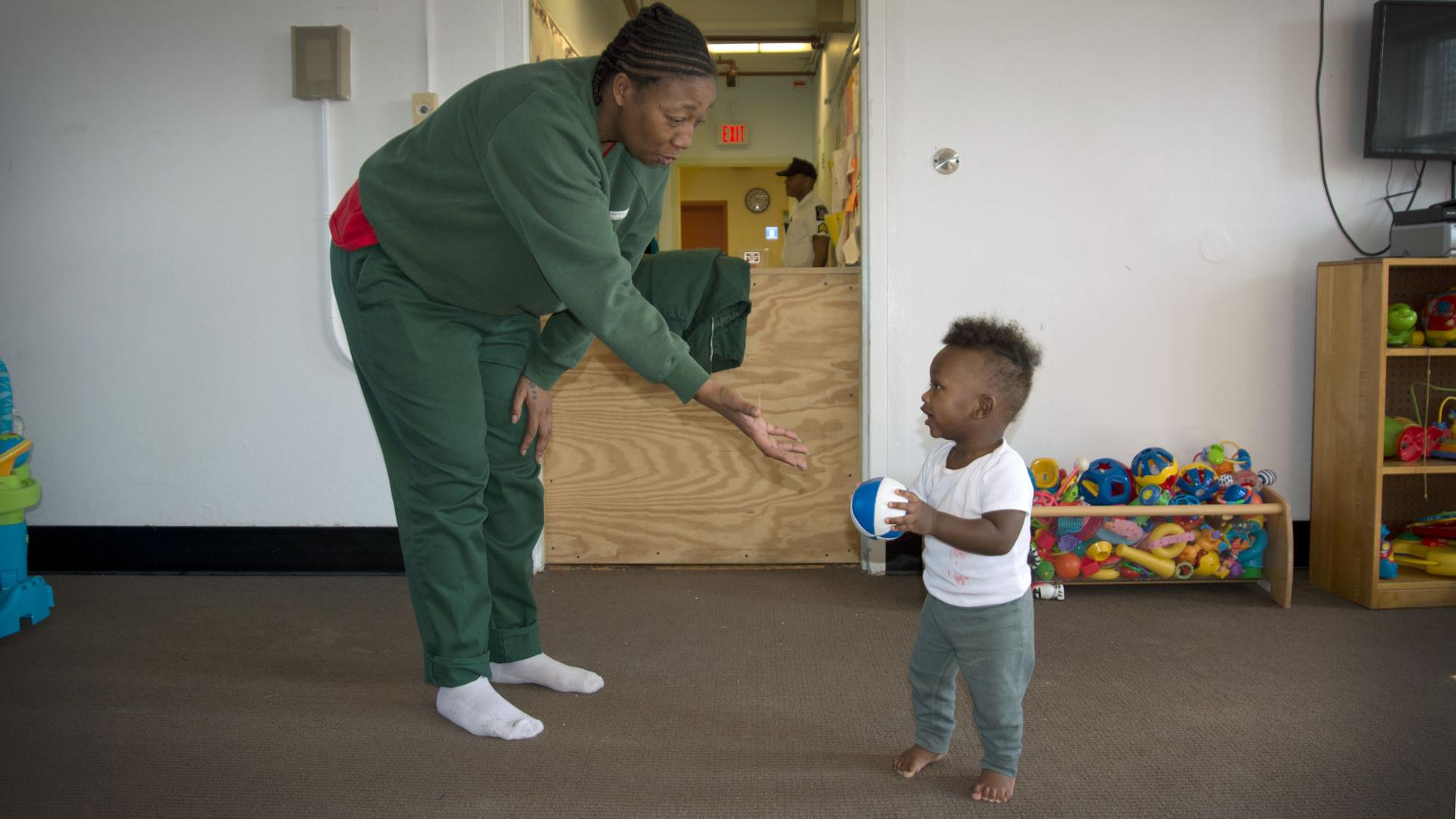 A woman bends over and reaches out a hand at a toddler, who is holding a ball. Behind him is a container of brightly colored children's toys.