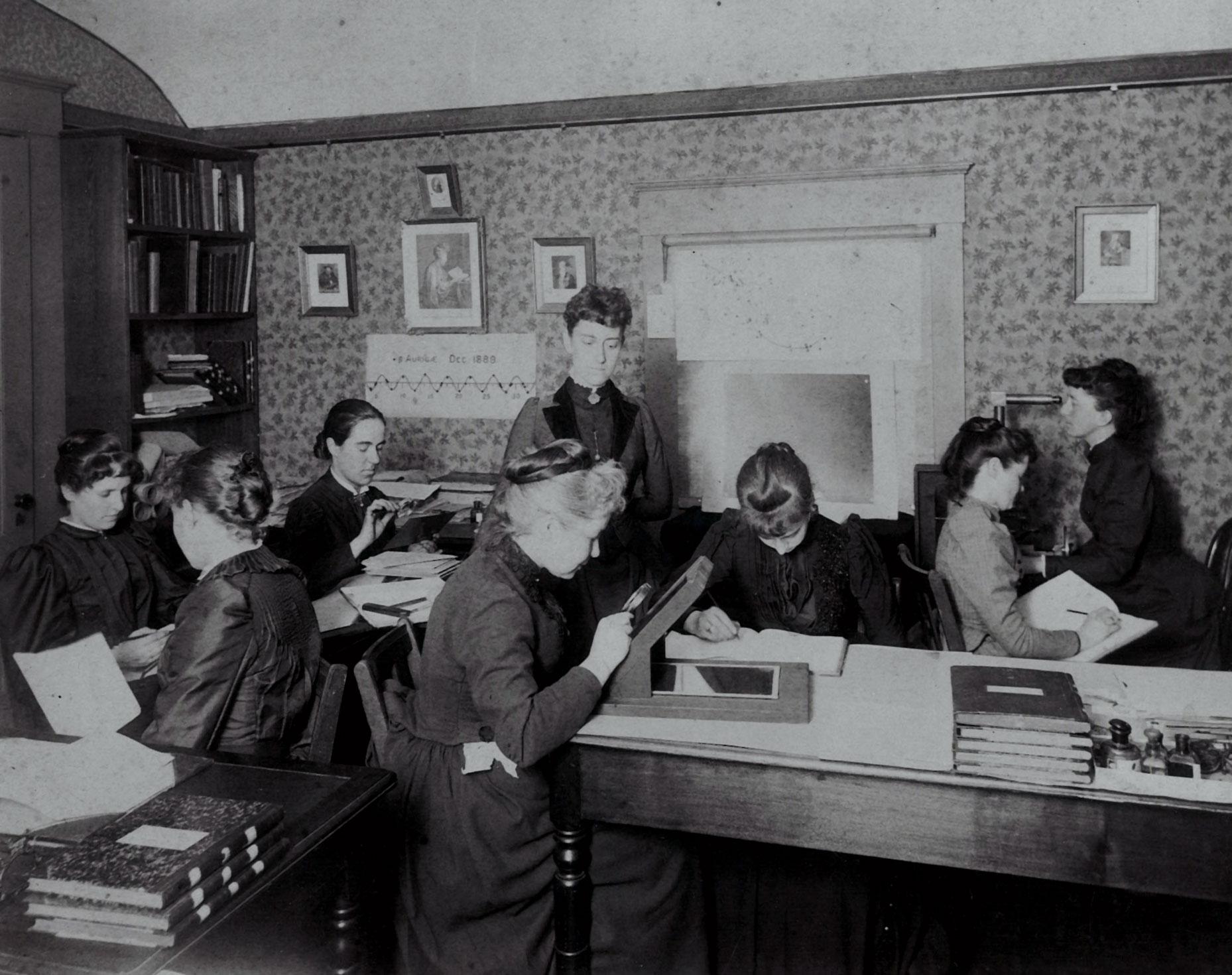 A group of women are sitting in a tiny room, some holding magnifying glasses, bent over boxes studying the glass plates. One stands in the center of the room, overseeing them. This is Williamina Fleming.