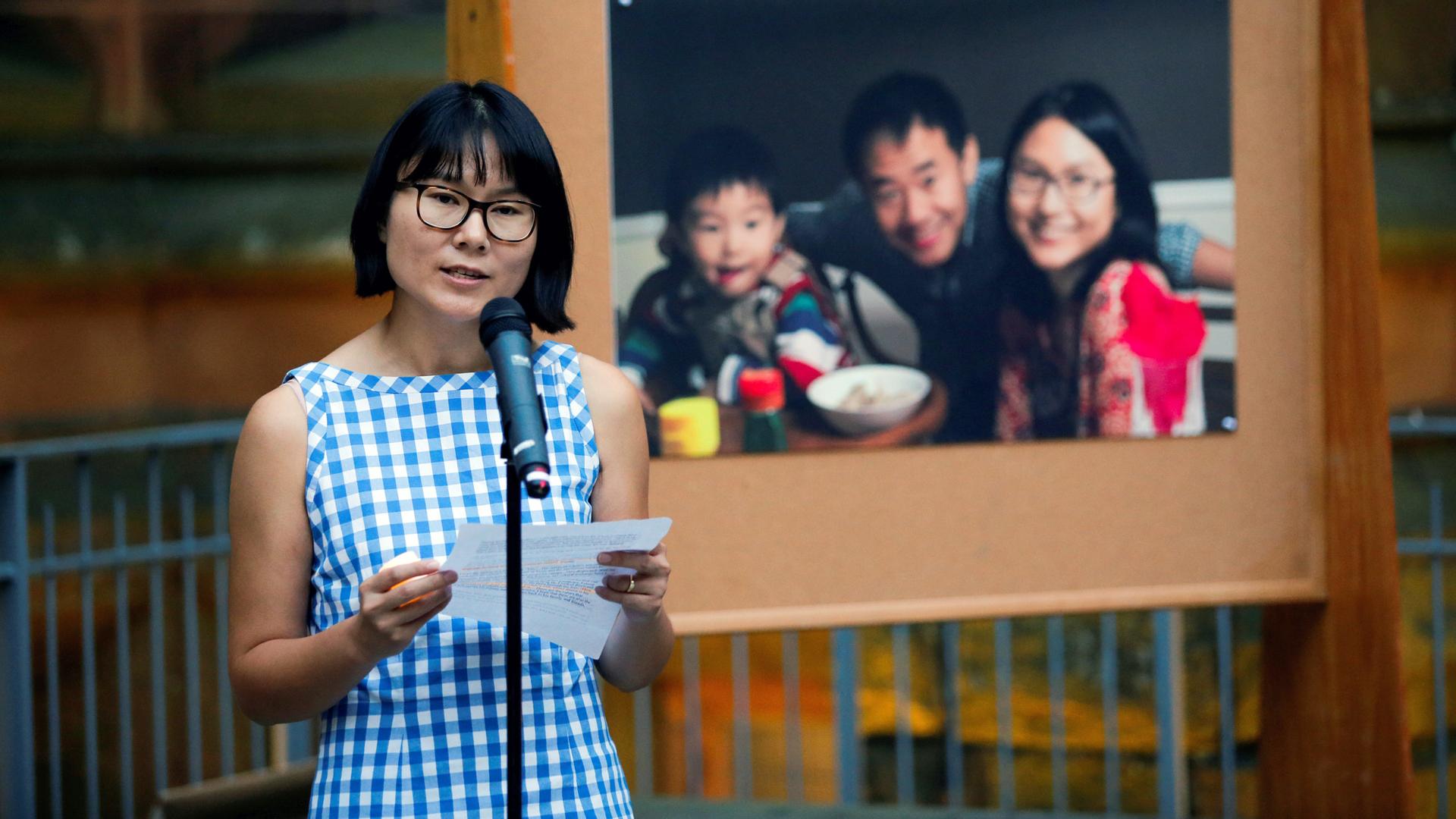 A woman speaks in front of a microphone. Behind her is a large image of her, with her husband and young son.