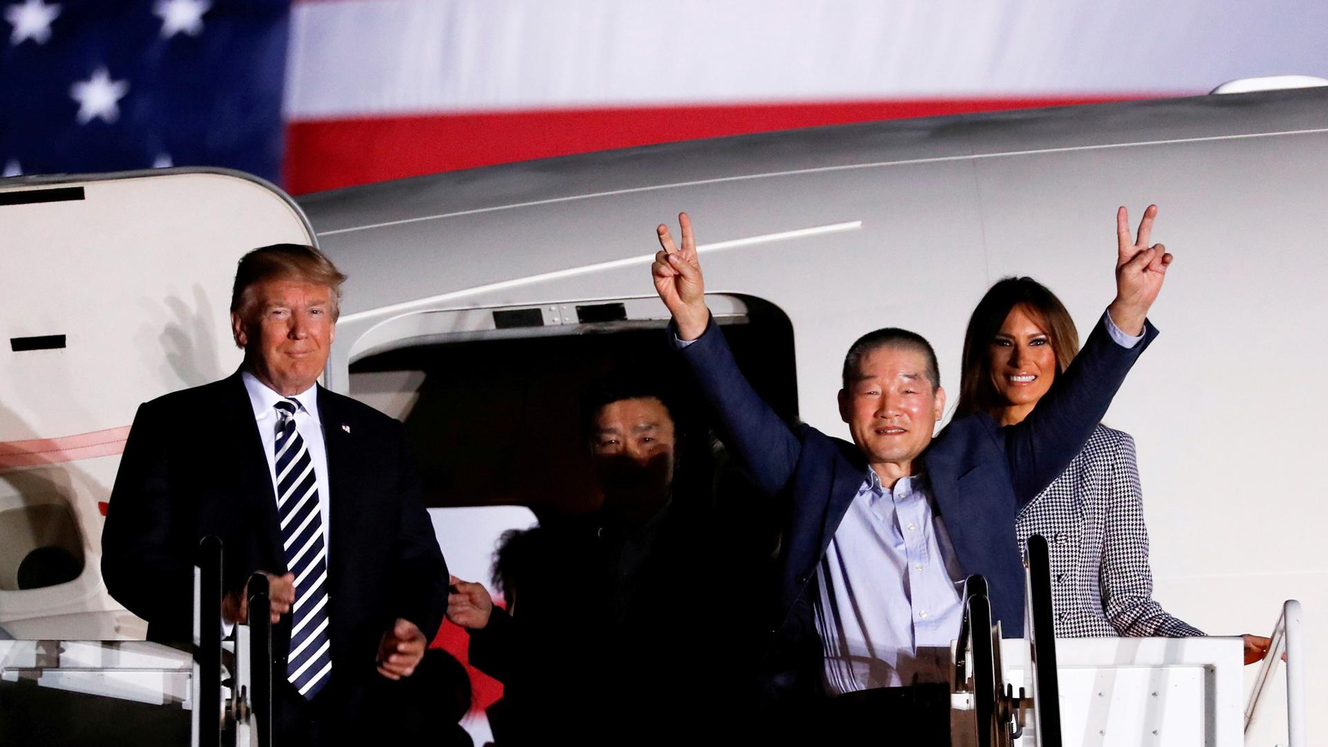 A man holds two fingers up on each hand as he exits an airplane. On the left is Donald Trump. Melania Trump is on the right.