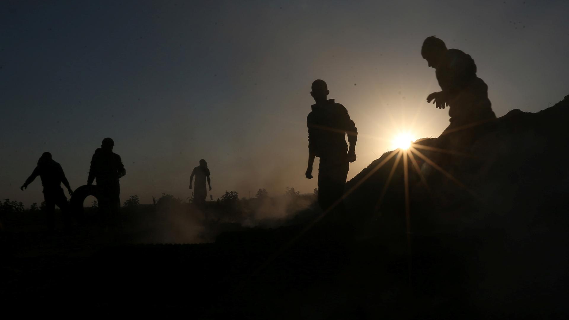 People are silhouetted against the sun as some smoke surrounds them. One man on the left rolls a tire.