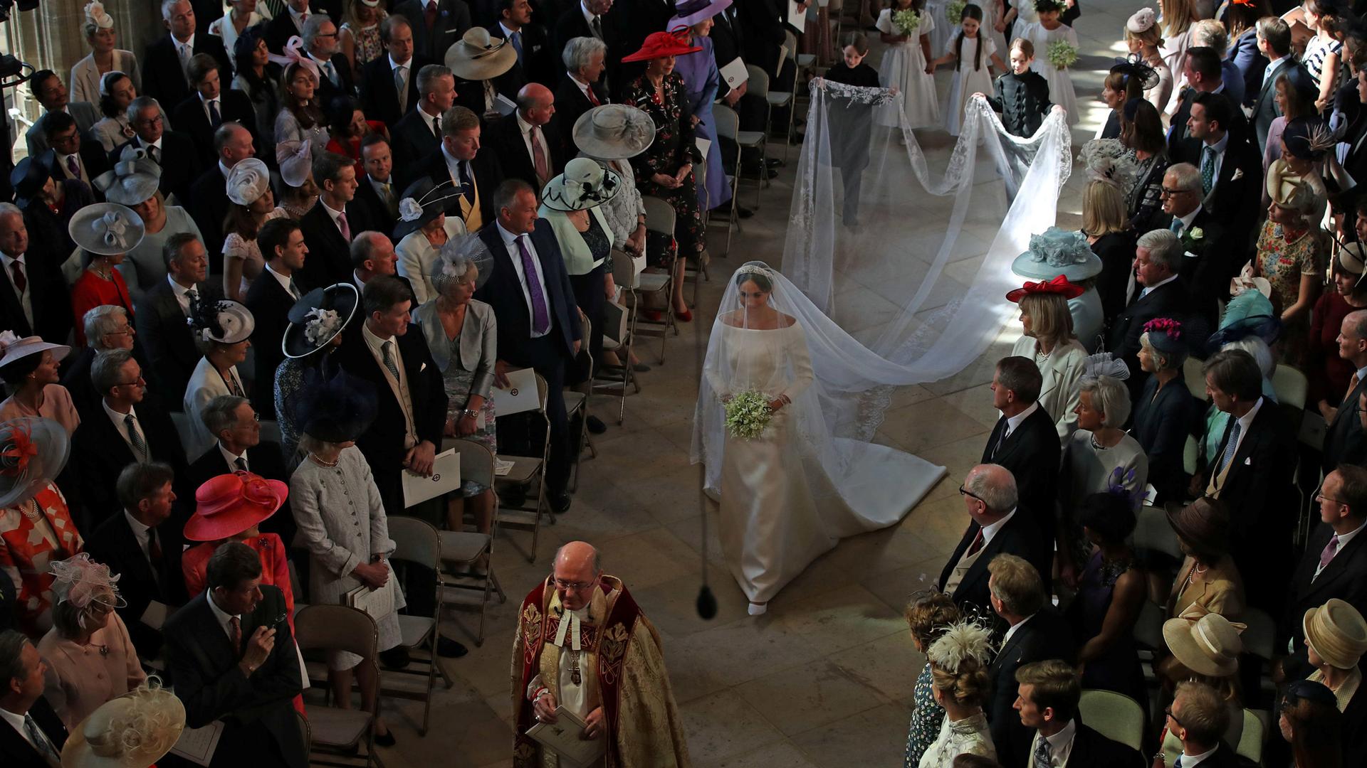 A bride, Meghan Markle, walks down the center of the aisle of a church. Her long white train drags beside her. Around her, everyone is standing. 