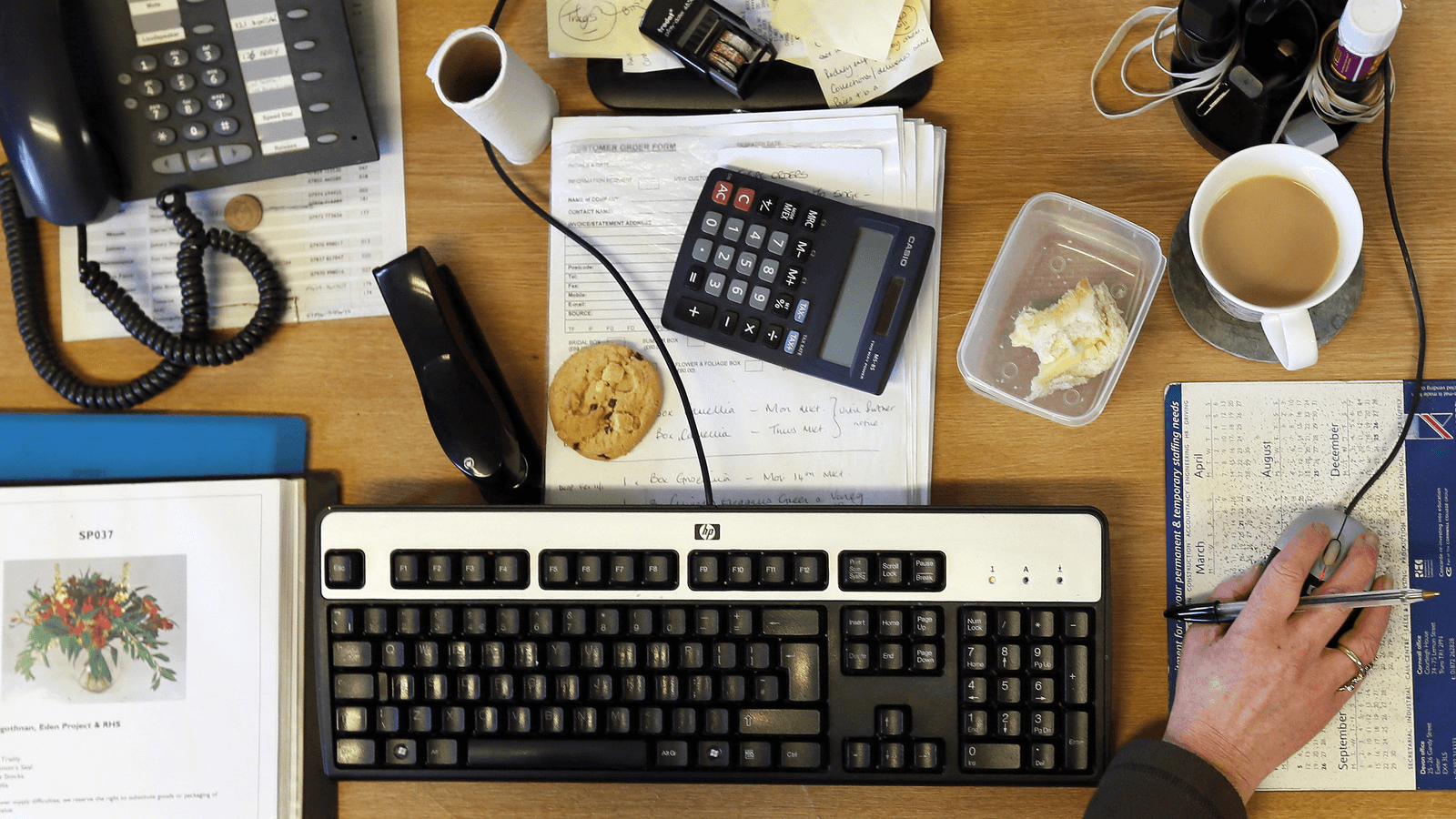 A desk full of food, keyboard, coffee and other stuff.