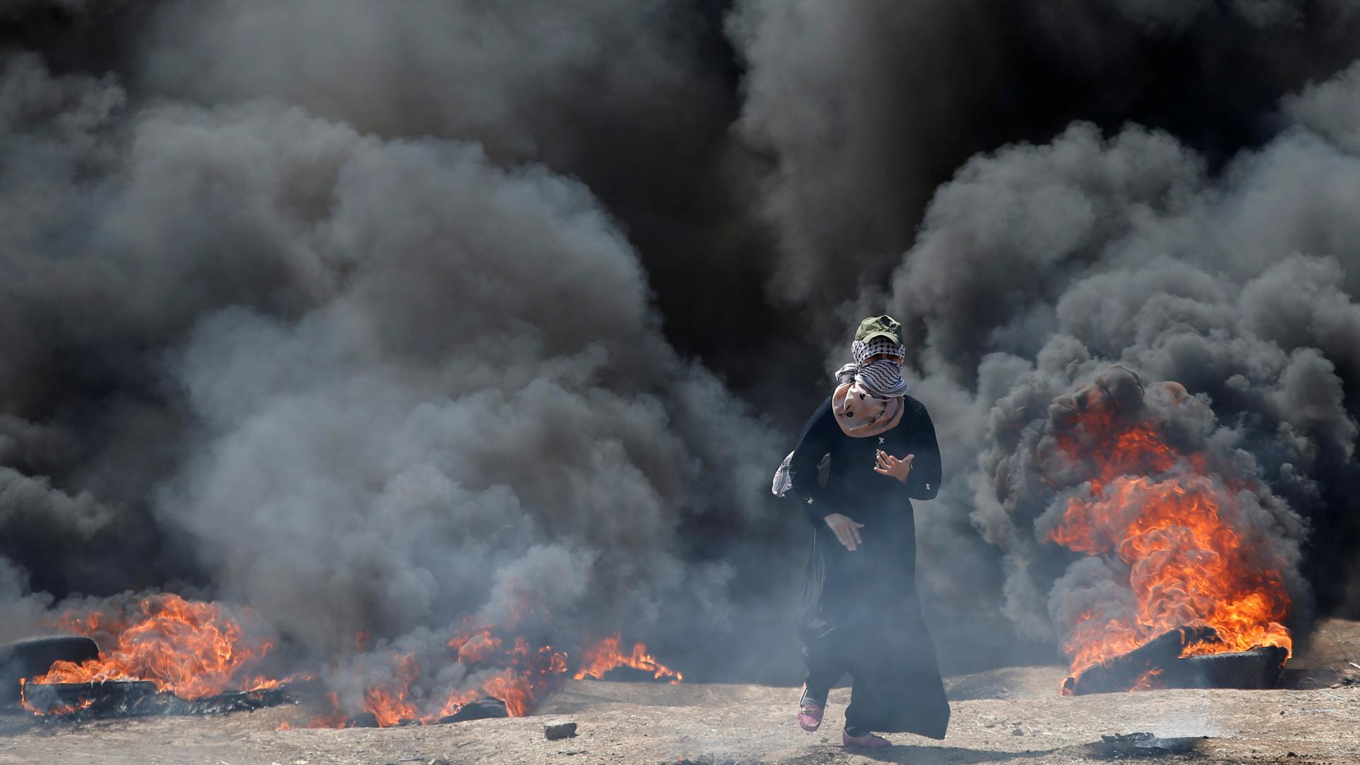 A Palestinian demonstrator walks with black smoke and tires burning in the background.
