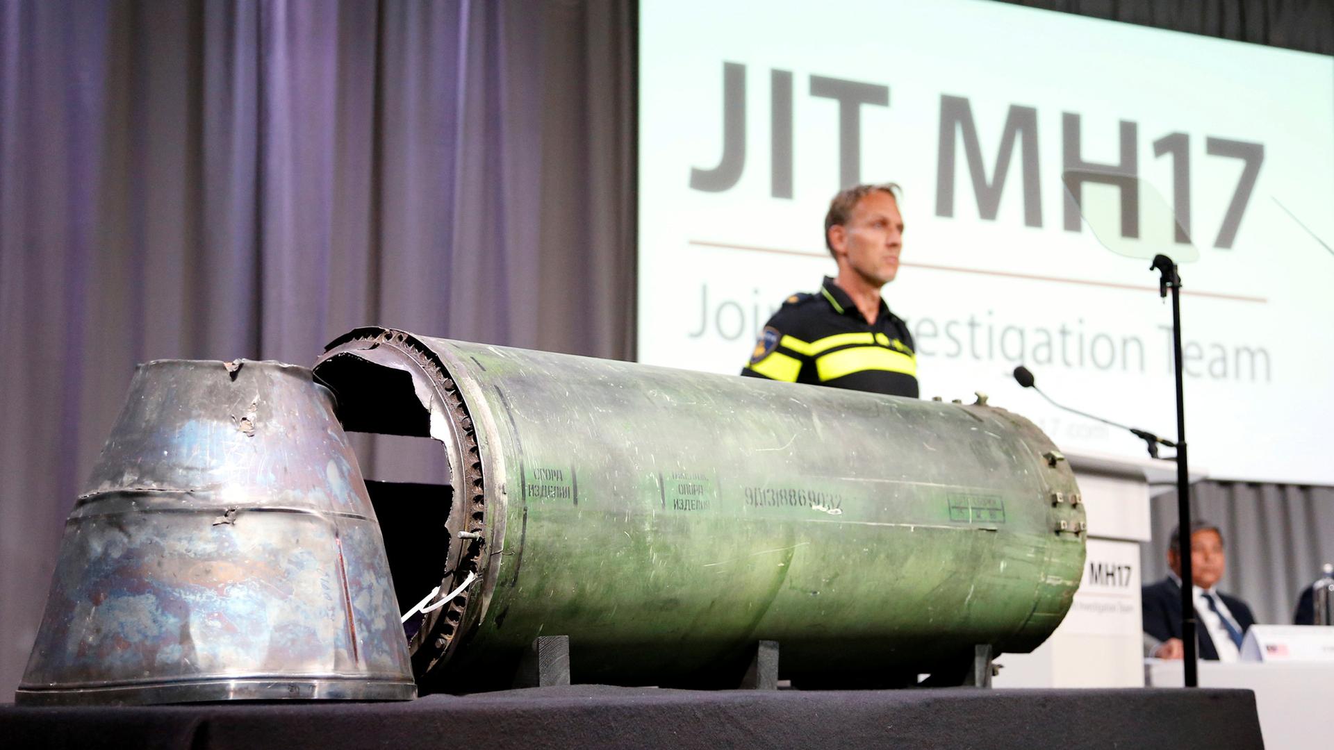 A large piece of silver meta, a damaged missile,l sits on a table while MH 17 is projected in the background of a screen.