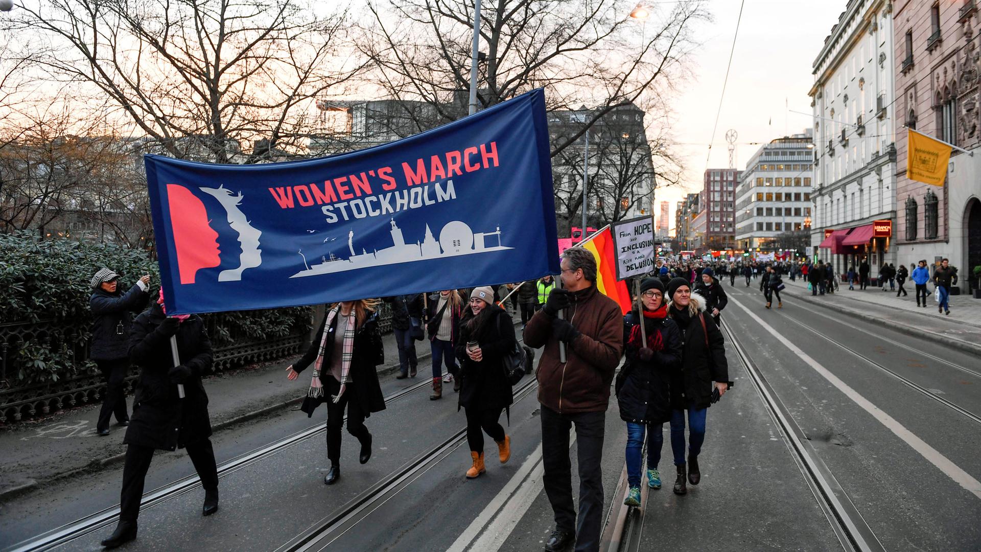 People carry a large banner that says "Women's March Stockholm" down a wide city street.