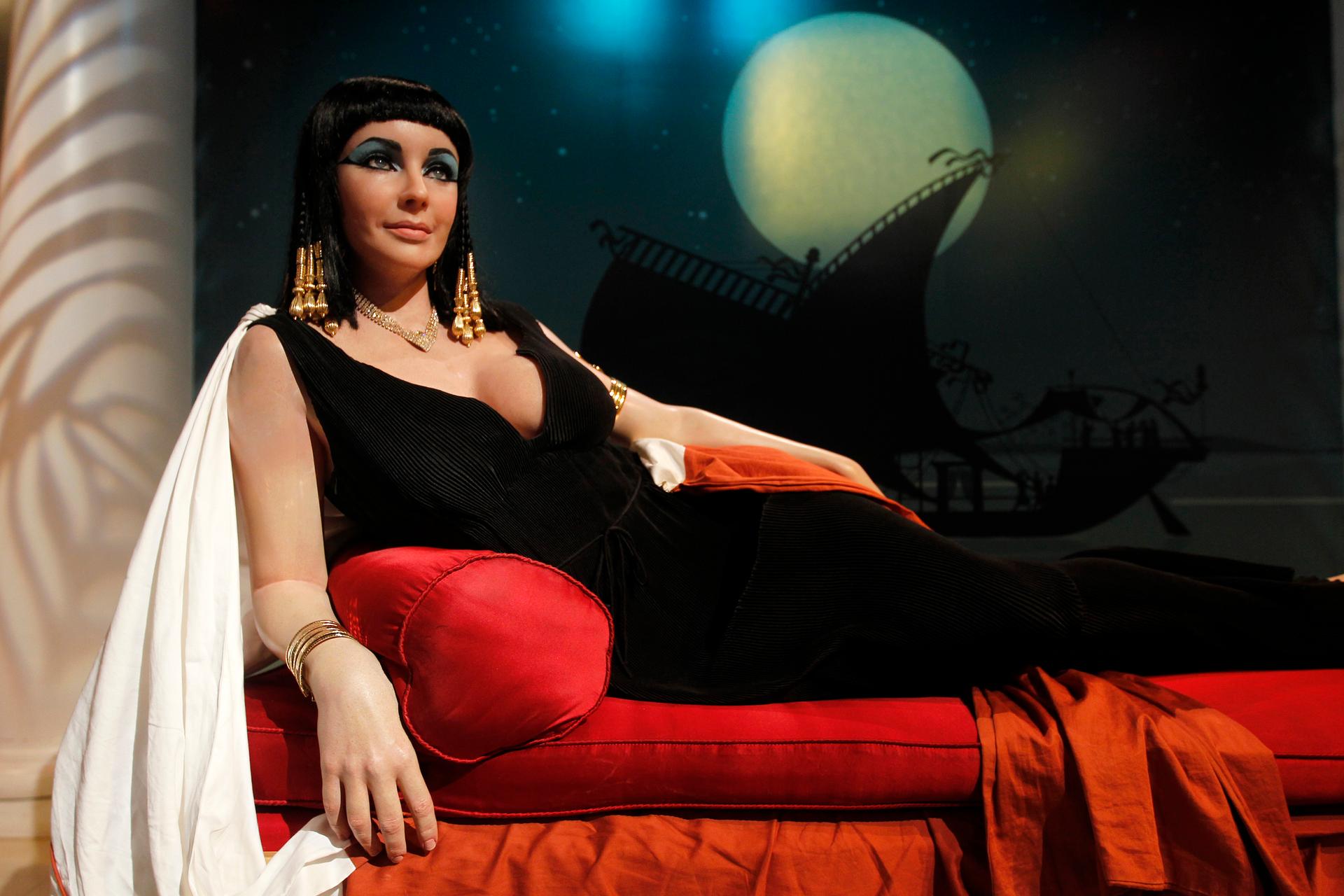 Elizabeth Tyler as Cleopatra lounges on a red cushion, he straight dark hair decorated with gold beads.