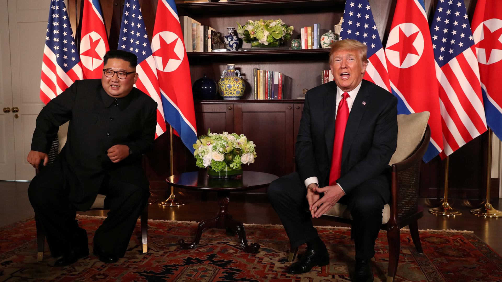 US President Donald Trump sits next to North Korea's leader Kim Jong-un with the flags of both countries behind them.