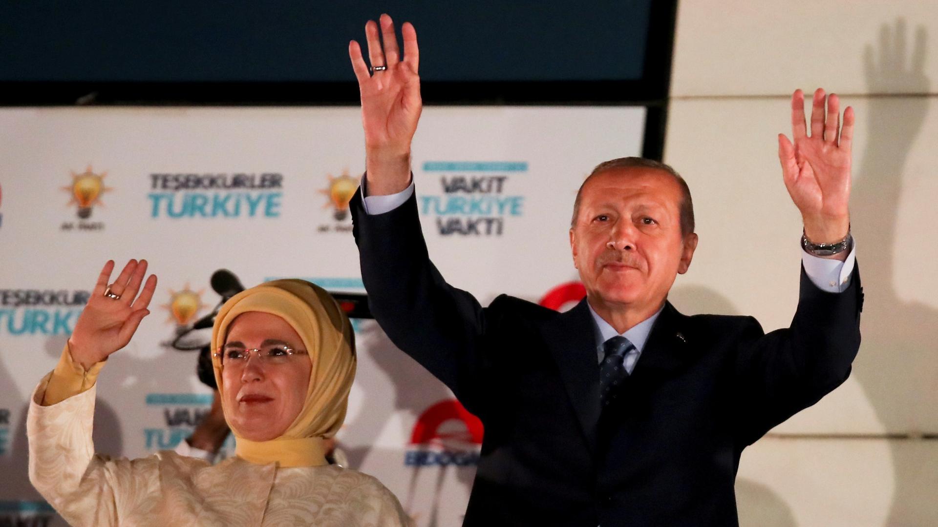 Turkish President Tayyip Erdoğan and his wife Emine Erdogan stand side-by-side with their arms up waving.