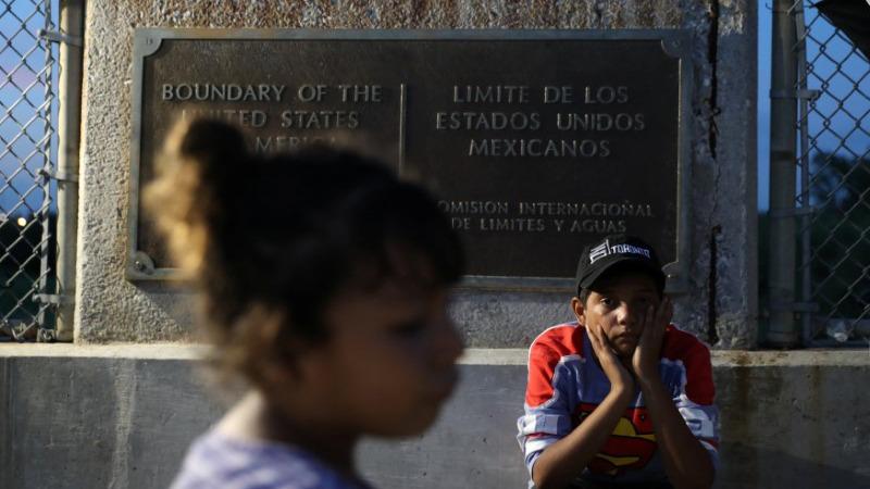 A young Honduran girl walks by in the near ground while a young boy in a Superman shirt and hat sits next to a sign noting the US border.