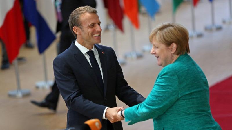 French President Emmanuel Macron, smiles as he warmly greets Germany's Chancellor Angela Merkel — the two holding hands.