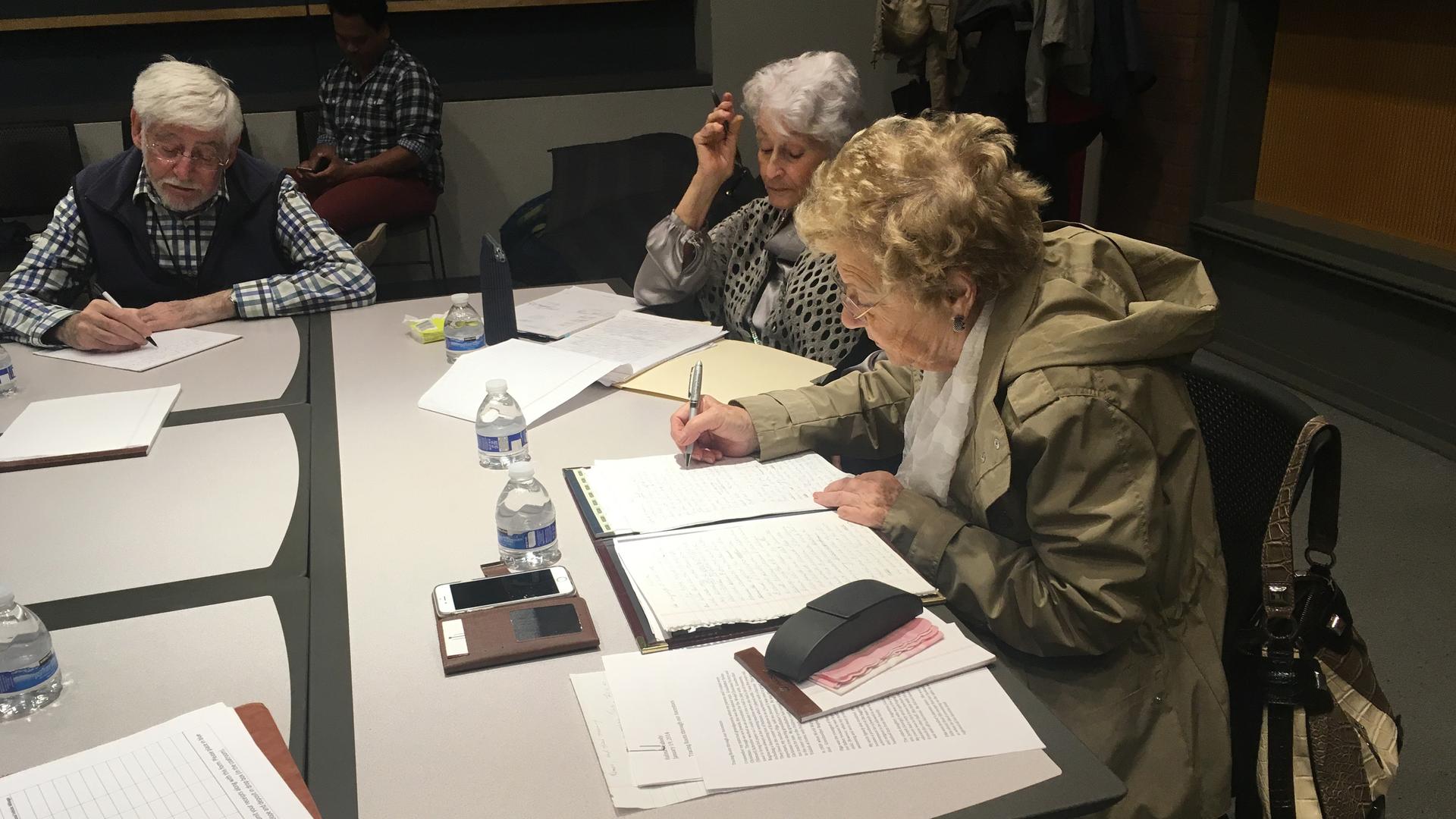 Three elderly Holocaust survivors sit at a table writing.