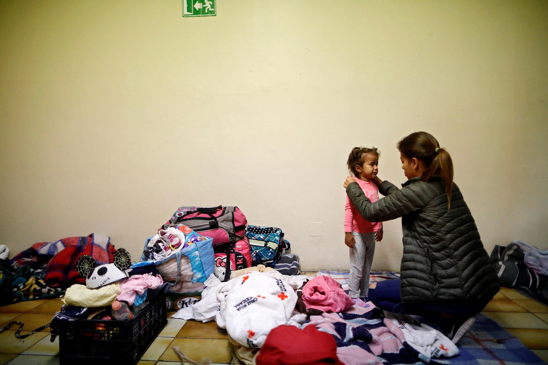 A woman dresses a girl at a shelter, surrounded by luggage and clothes